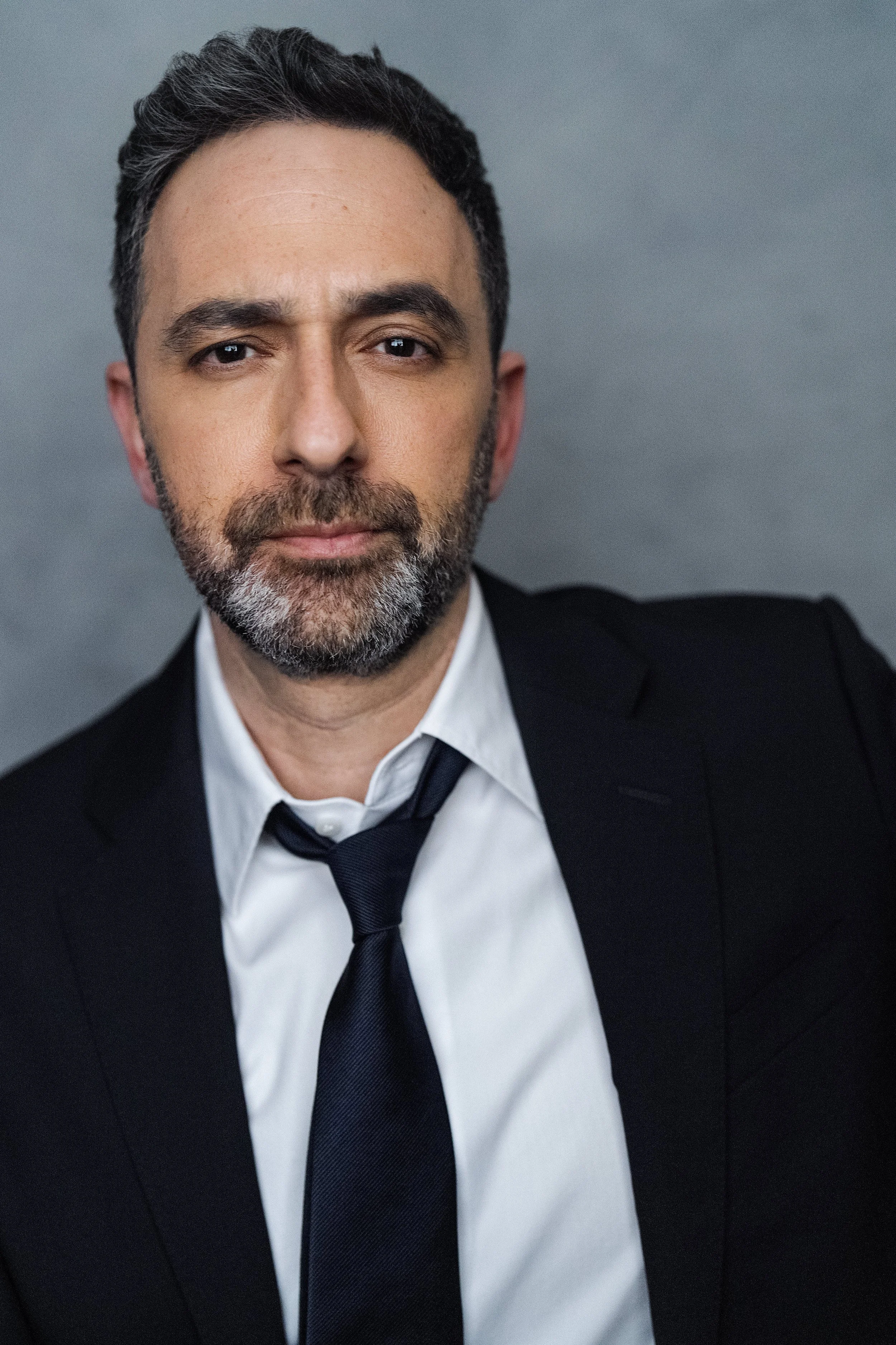 A middle-aged man with dark hair, beard, and mustache, wearing a black suit, white shirt, and black tie, looking directly at the camera with a serious expression.