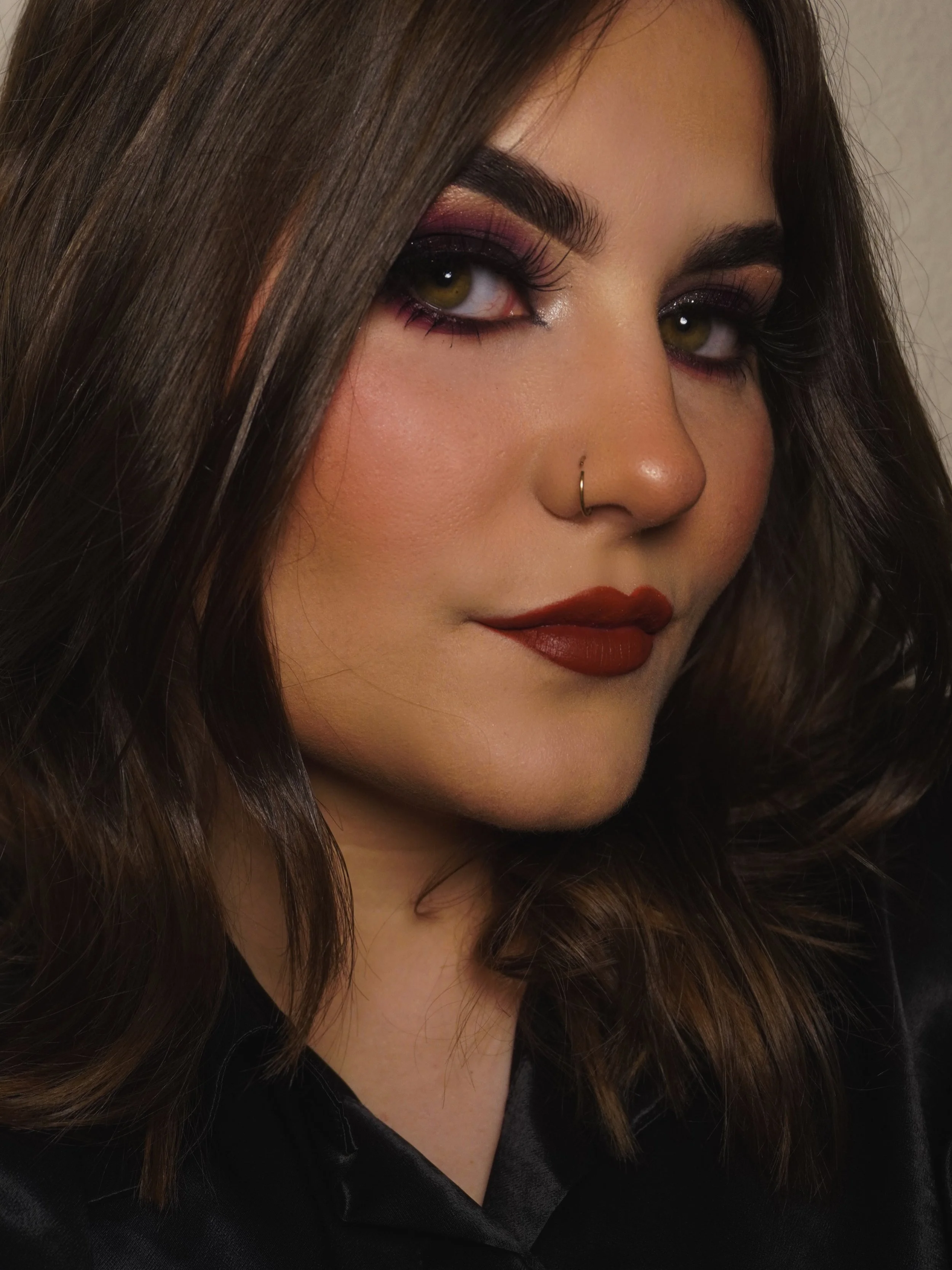 Close-up of a woman with dark brown hair, wearing makeup including pink and purple eyeshadow, false eyelashes, and dark red lipstick. She has a small gold nose ring.