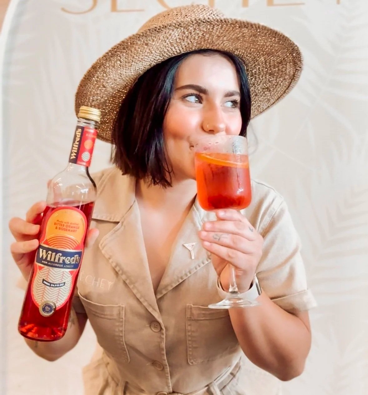 A woman wearing a straw hat and beige shirt holding a bottle of Wilfred's orange and rosehip flavored sparkling beverage in one hand and a glass filled with a red cocktail garnished with an orange slice in the other. She is smiling and looking to her right.