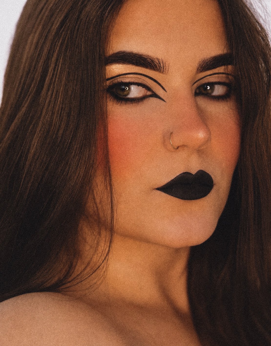 Close-up of a woman with dark brown hair, bold black makeup, black lipstick, and a nose ring, winking with makeup details around her eyes.