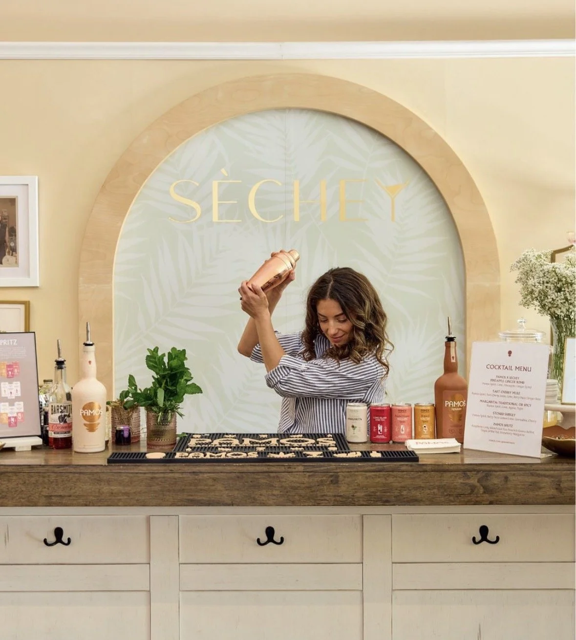 A woman bartending at a bar with a large circular mirror behind her that says 'SÉCHÉ'. She is pouring a drink into a copper shaker, surrounded by various bottles, cans, and a small potted plant on a wooden bar counter.