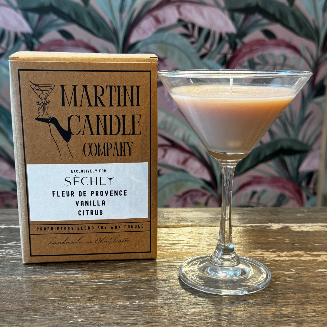 A cocktail glass filled with a pinkish-orange drink, placed on a wooden surface next to a box of Martini Candle Company soy wax candle, with a floral wallpaper background.