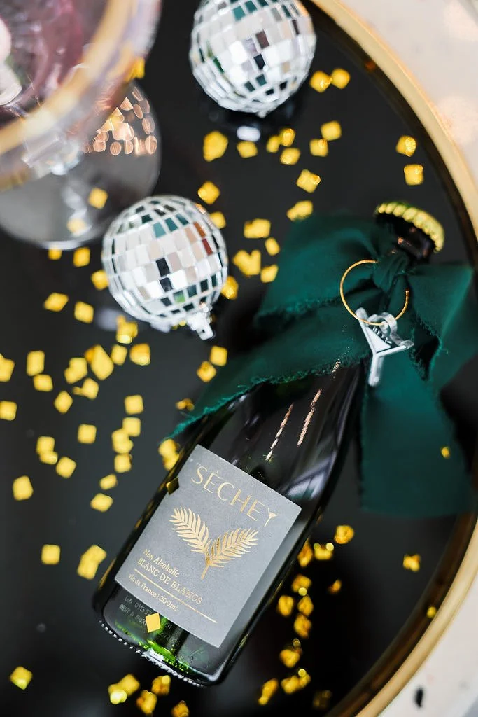 A black sparkling wine bottle with a green ribbon, surrounded by yellow confetti and two disco balls, on a black surface.