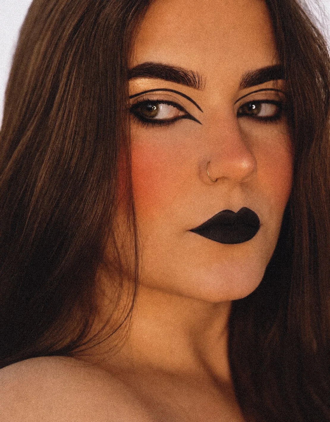 Close-up of a woman with makeup, dark lipstick, bold eyeliner, and a septum piercing.