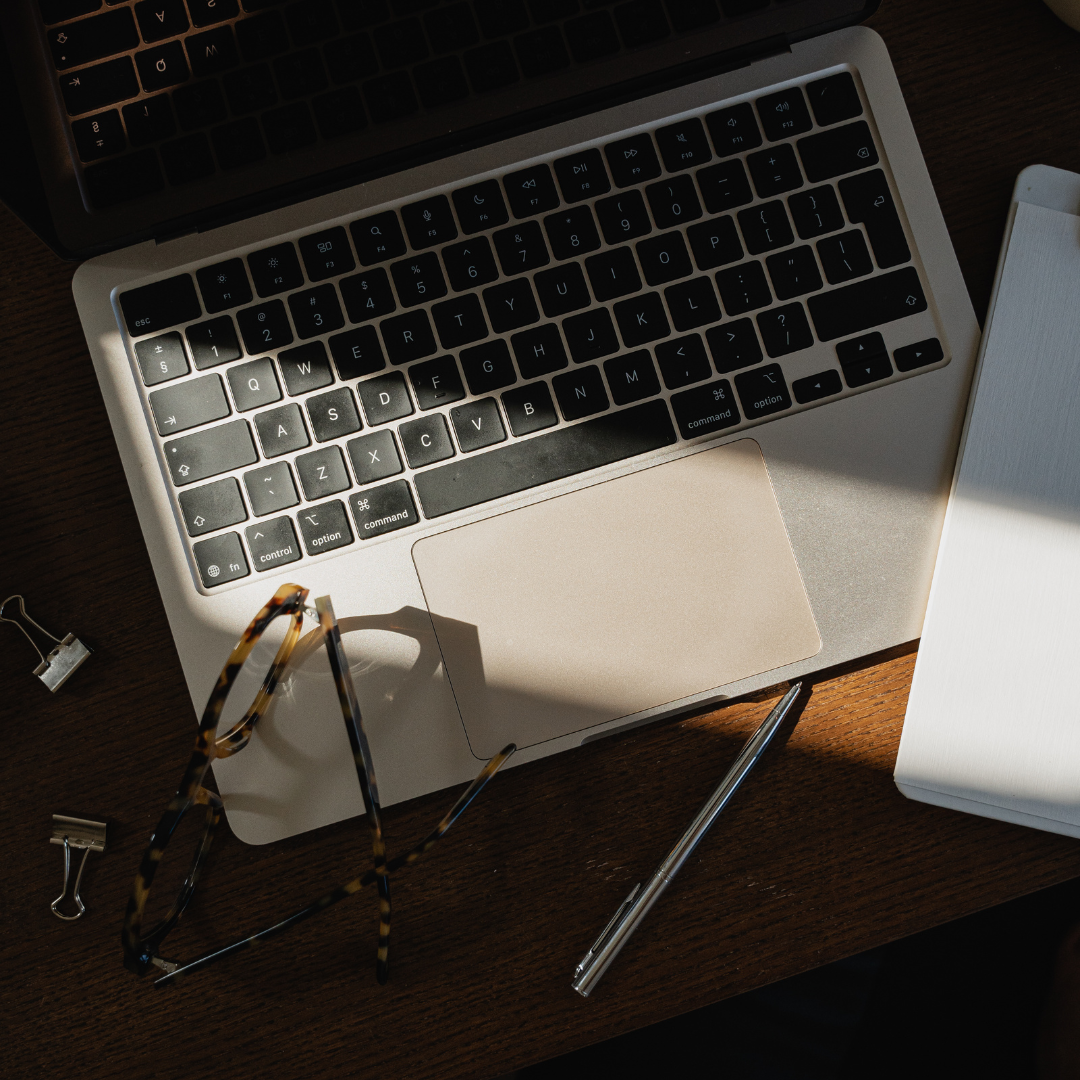 Top-down view of a laptop on a wooden desk, with a pair of tortoiseshell glasses, a pen, two binder clips, and a closed white notebook nearby.