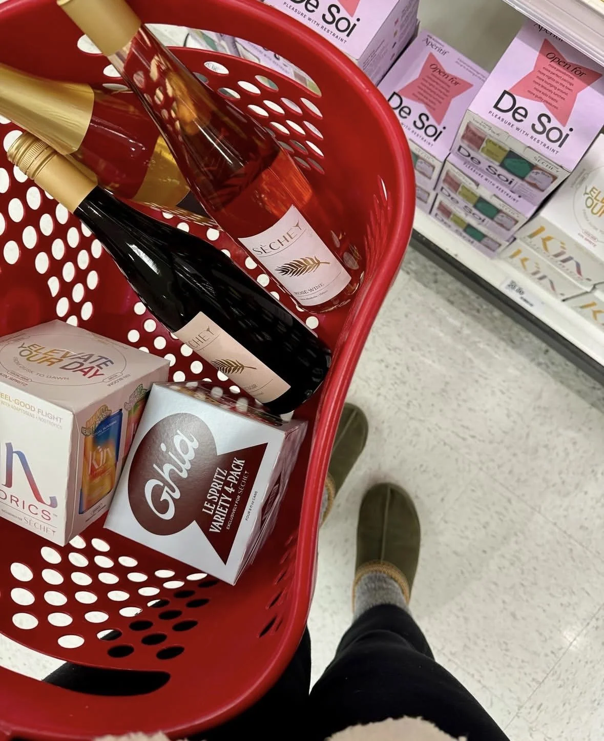 A red shopping basket containing wine bottles and snack boxes, with a person wearing dark pants and brown shoes standing nearby in a store aisle.