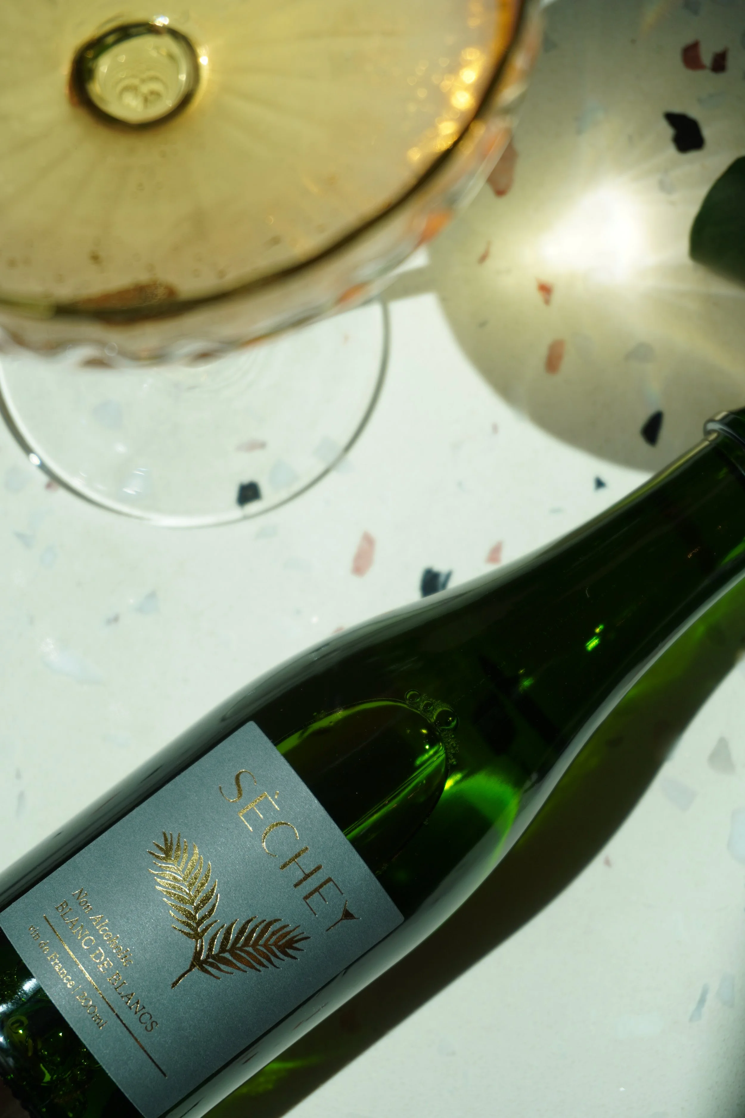 A green bottle of Secateurs wine lying on a white speckled surface next to a clear wine glass and a plant with a blurred background.