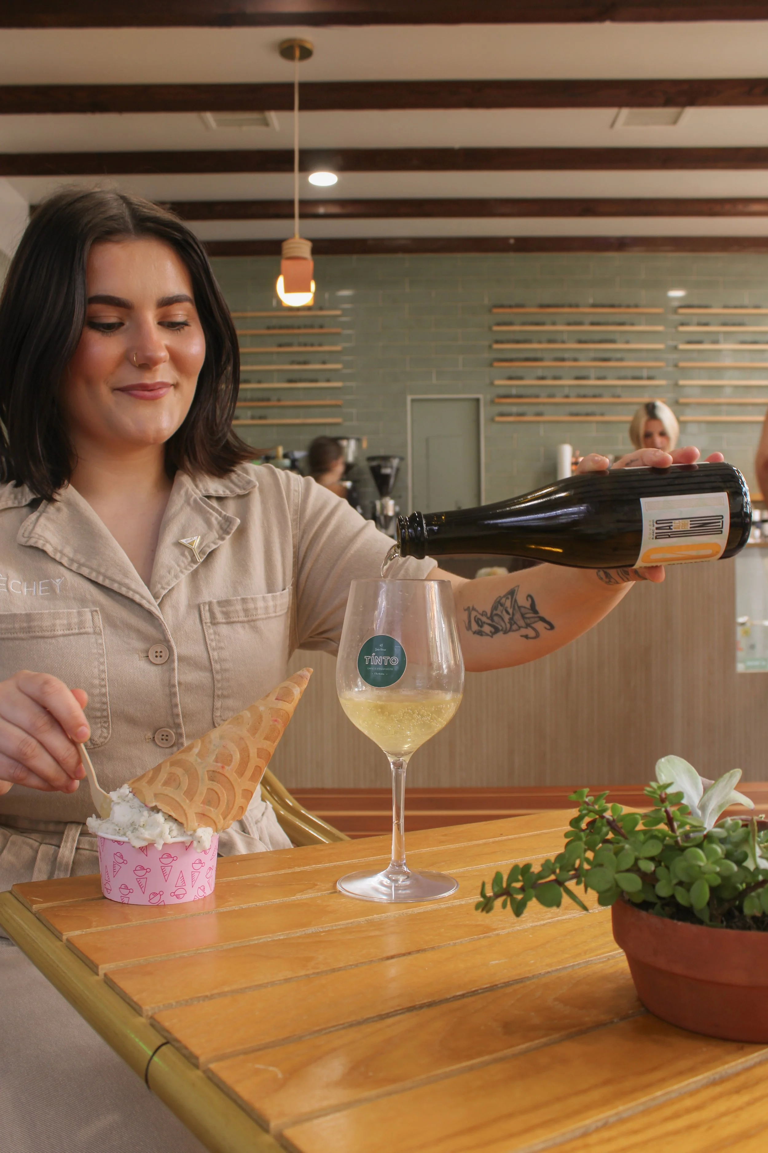 A woman with shoulder-length dark hair and a nose piercing pours white wine into a glass at a wooden table. There is a cup with ice cream and an ice cream cone nearby, a potted plant, and people in the background.
