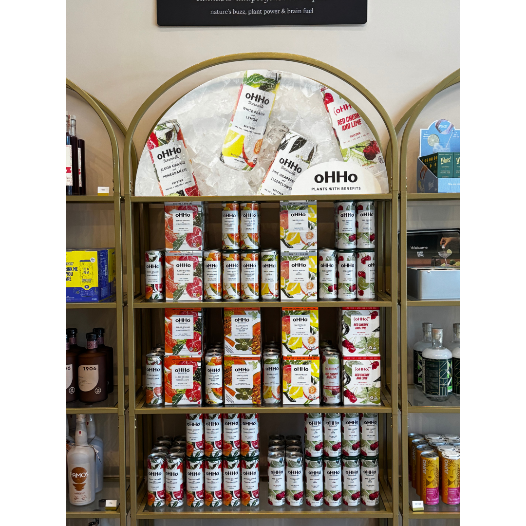 A display shelf filled with cans and cartons of oHHo flavored sparkling water, featuring various fruit flavors such as lemon, cherry, lime, and blood orange. The cans and cartons are arranged neatly on the gold-colored shelves with a backdrop of a sign promoting the drinks.