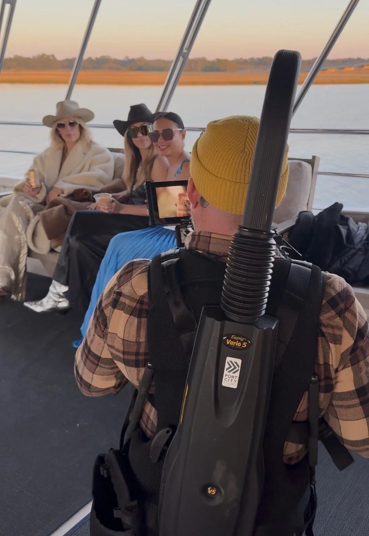 Four women sitting on a boat, two wearing hats and sunglasses, holding drinks, with a man filming them from behind. The boat has a view of water and a distant shoreline with trees during sunset.