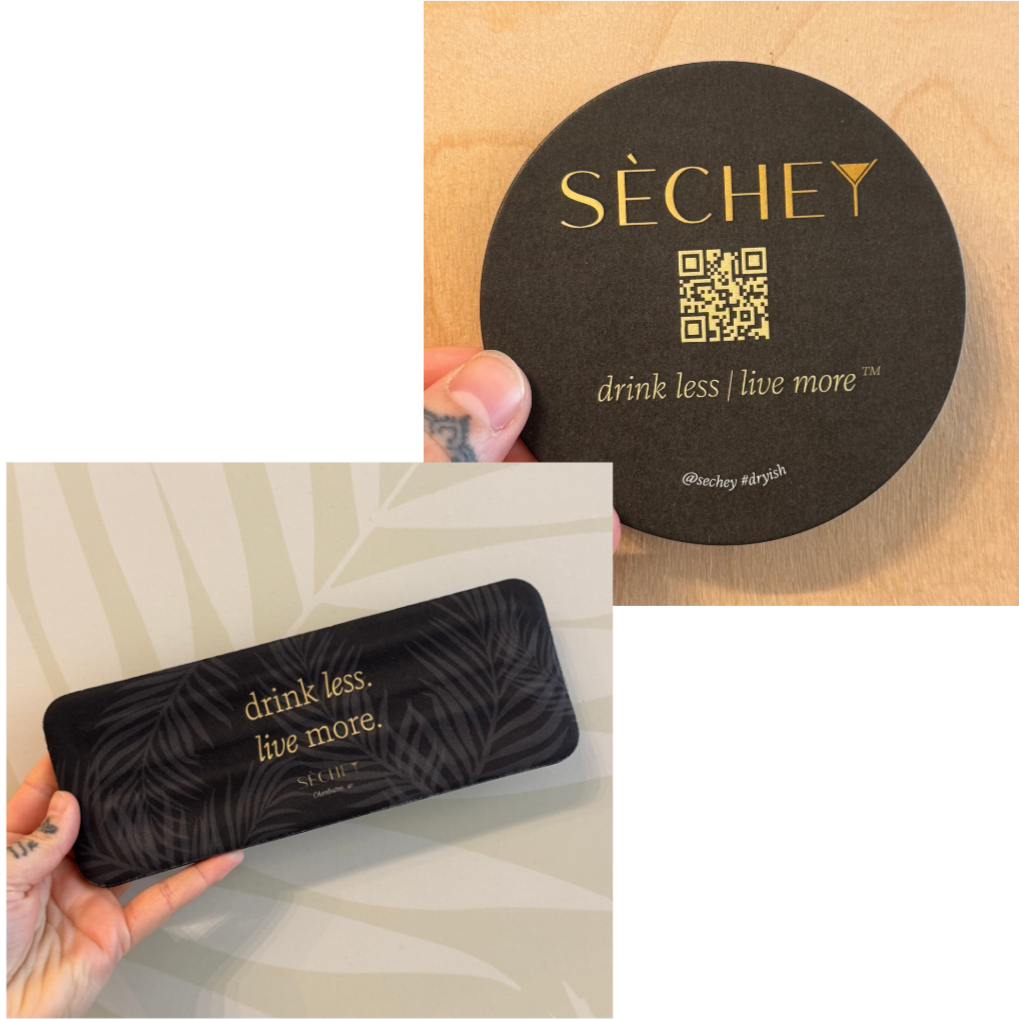 Two black items with the slogan "drink less. live more" in gold lettering, one round coaster and one rectangular case, held by a person with tattoos on their fingers.