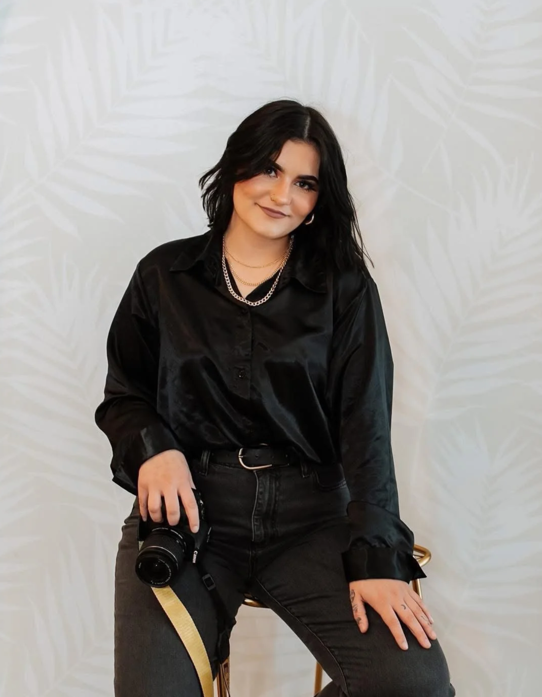 A young woman with short black hair, wearing a black satin shirt, black jeans, silver jewelry, and holding a camera, sitting on a gold stool against a white wall with a subtle leaf pattern.