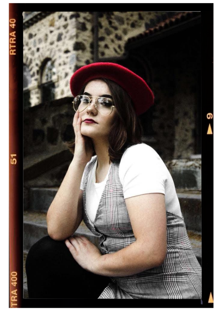 A young woman sitting outdoors on stone steps, wearing a red beret, glasses, a white t-shirt, and a plaid dress, resting her chin on her hand and looking at the camera.