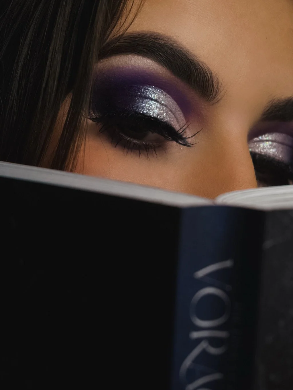 Close-up of a woman's eye with purple and silver glitter eyeshadow, visible eyelashes, and dark eyebrows, partially obscured by a book.