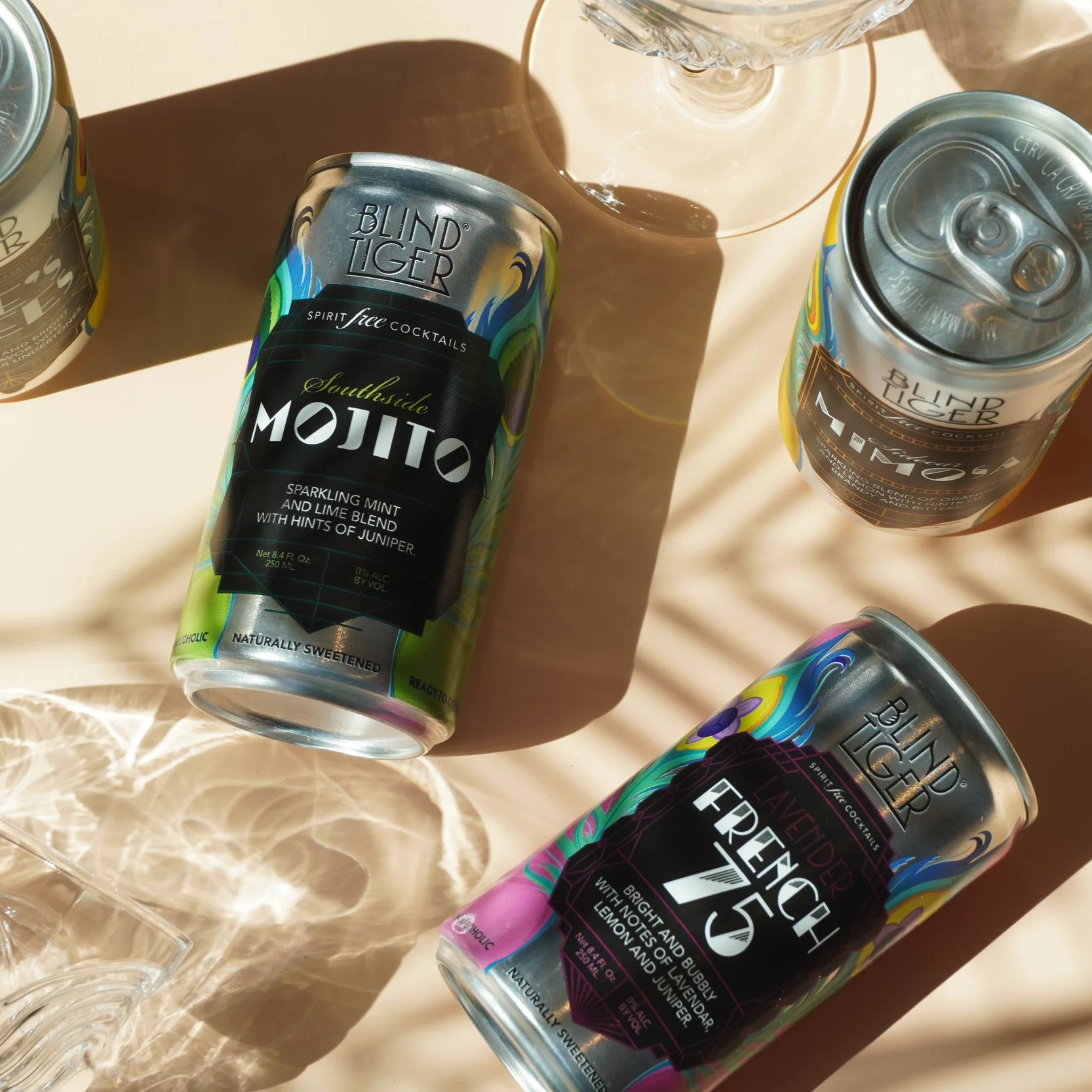 Several cans of Blind Tiger alcohol-free cocktail drinks and a glass on a tan surface with sunlight creating shadows.