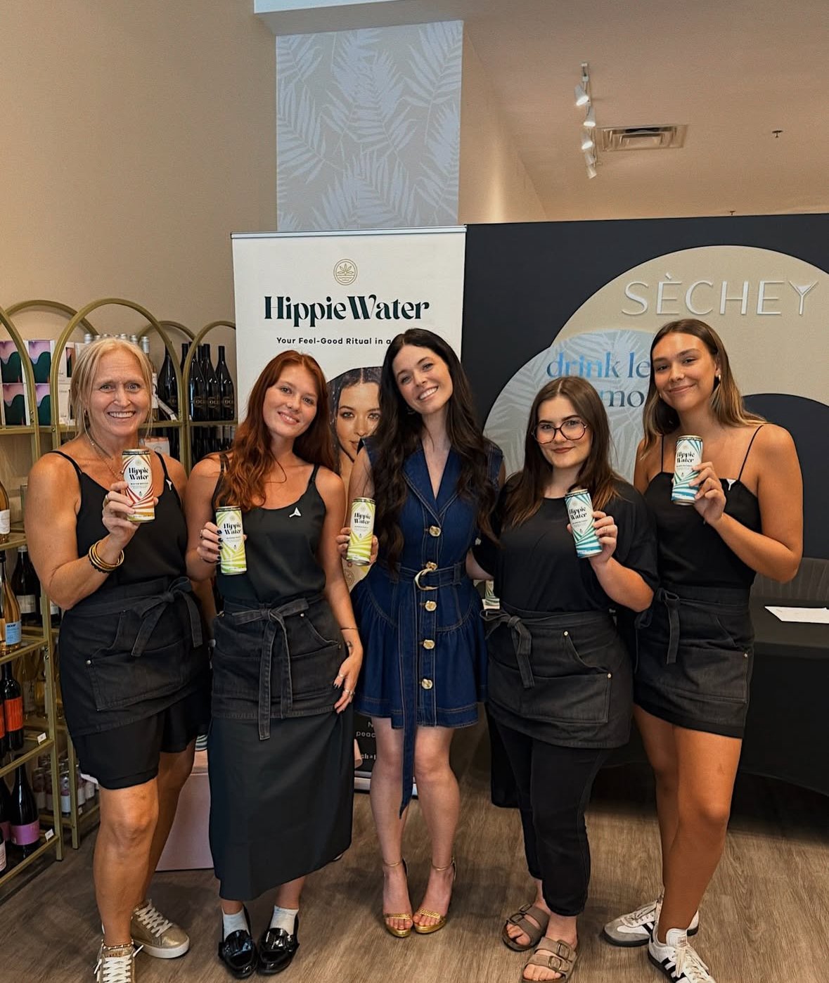 Five women at a promotional event holding cans of Hippie Water, standing in front of banners and shelves with wine bottles, dressed in black or blue outfits.