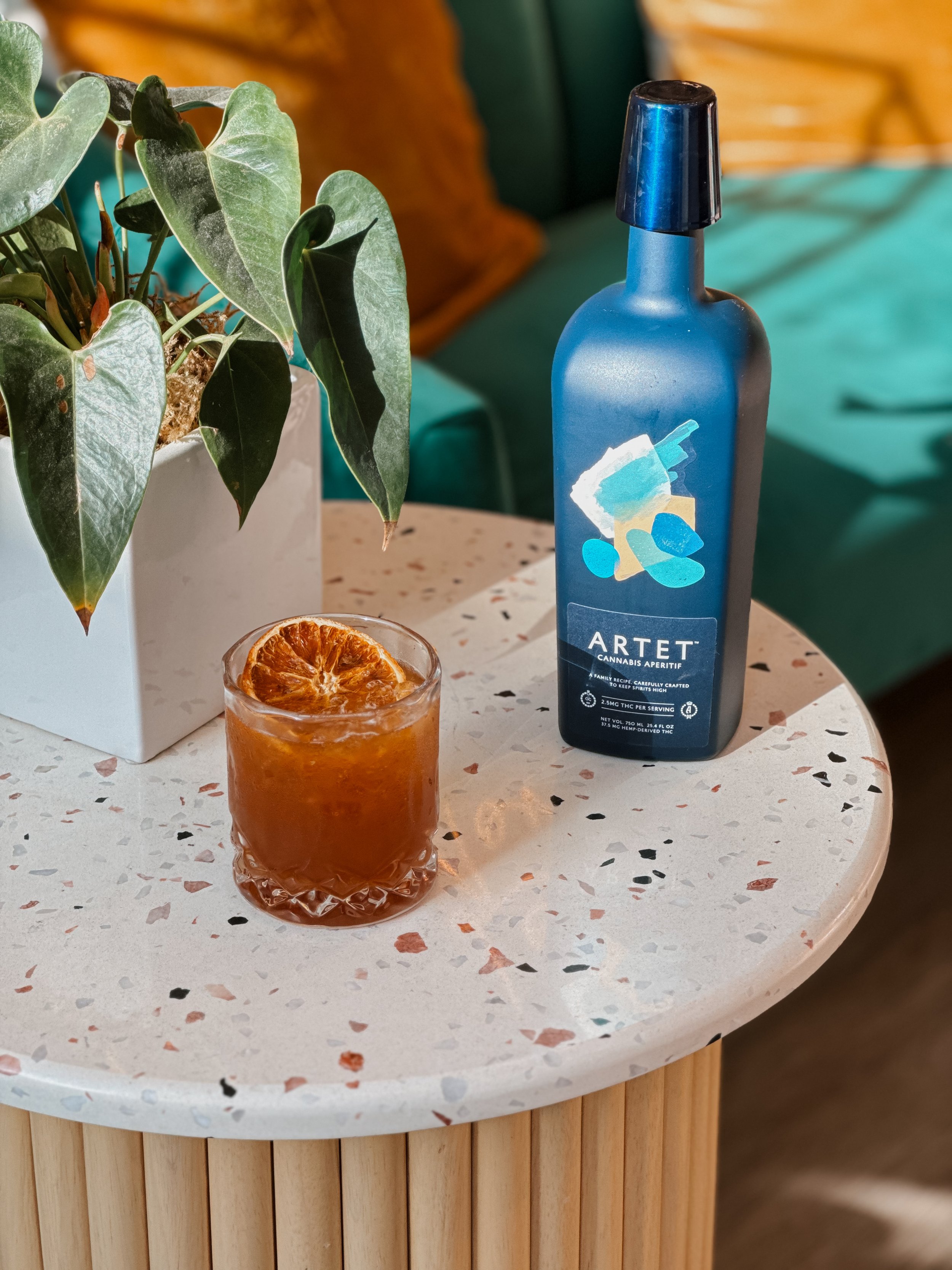 A glass of iced tea with a slice of dried orange on top, a potted plant with green leaves, and a blue cannabis-infused beverage bottle on a terrazzo table with a speckled pattern, in a cozy indoor setting.
