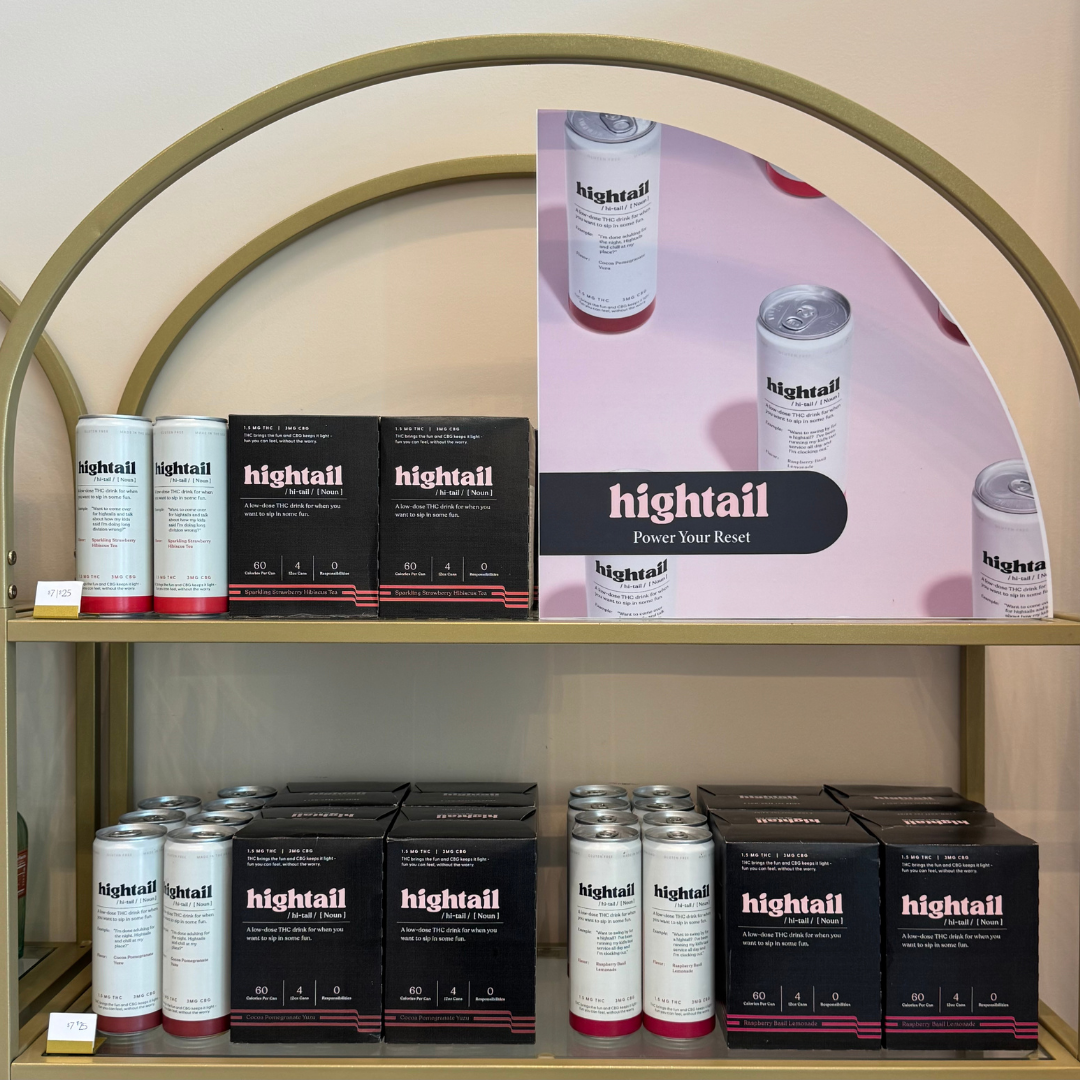 Display shelf with cans and boxes of hightail THC drink products, alongside a promotional poster for hightail with product bottles on a pink background.