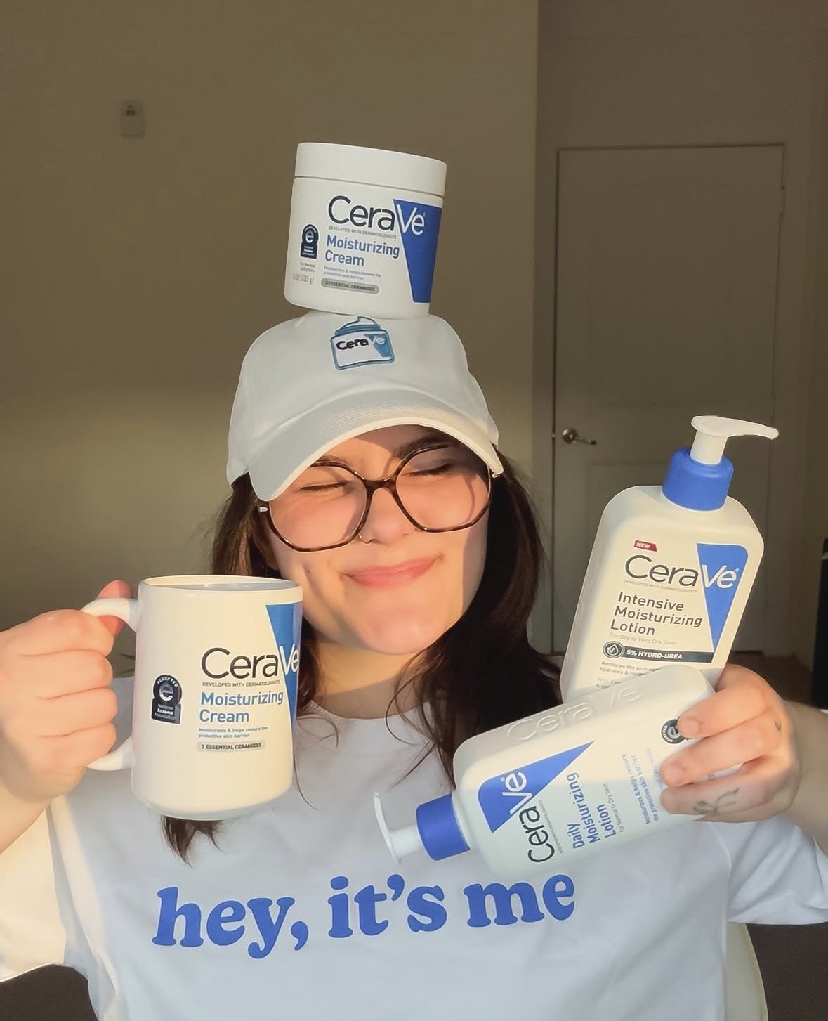 A woman smiling with her eyes closed, wearing glasses and a white cap, holding a coffee mug and a bottle of CeraVe moisturizing lotion, with another CeraVe Moisturizing Cream container balanced on her head.