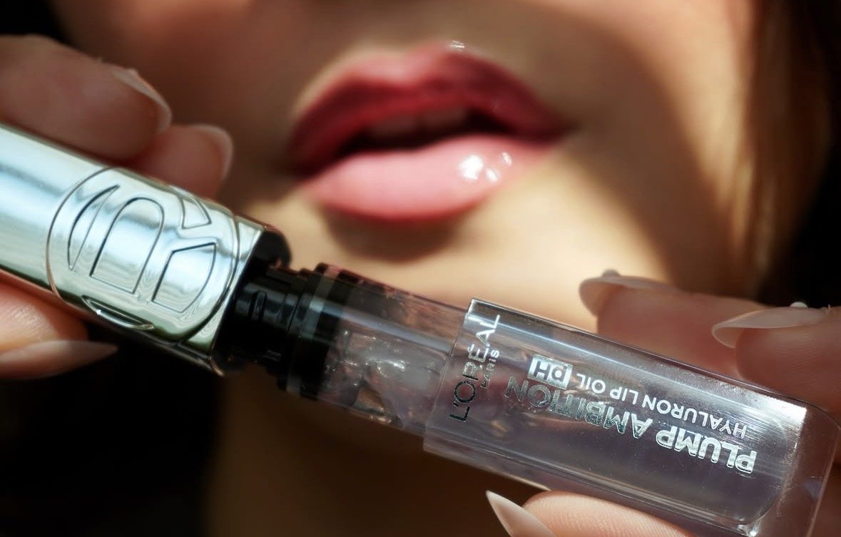 Close-up of a person applying lip gloss with a tube labeled 'Plump Amplification Hydrating Lip Oil' by L'Oréal Paris.