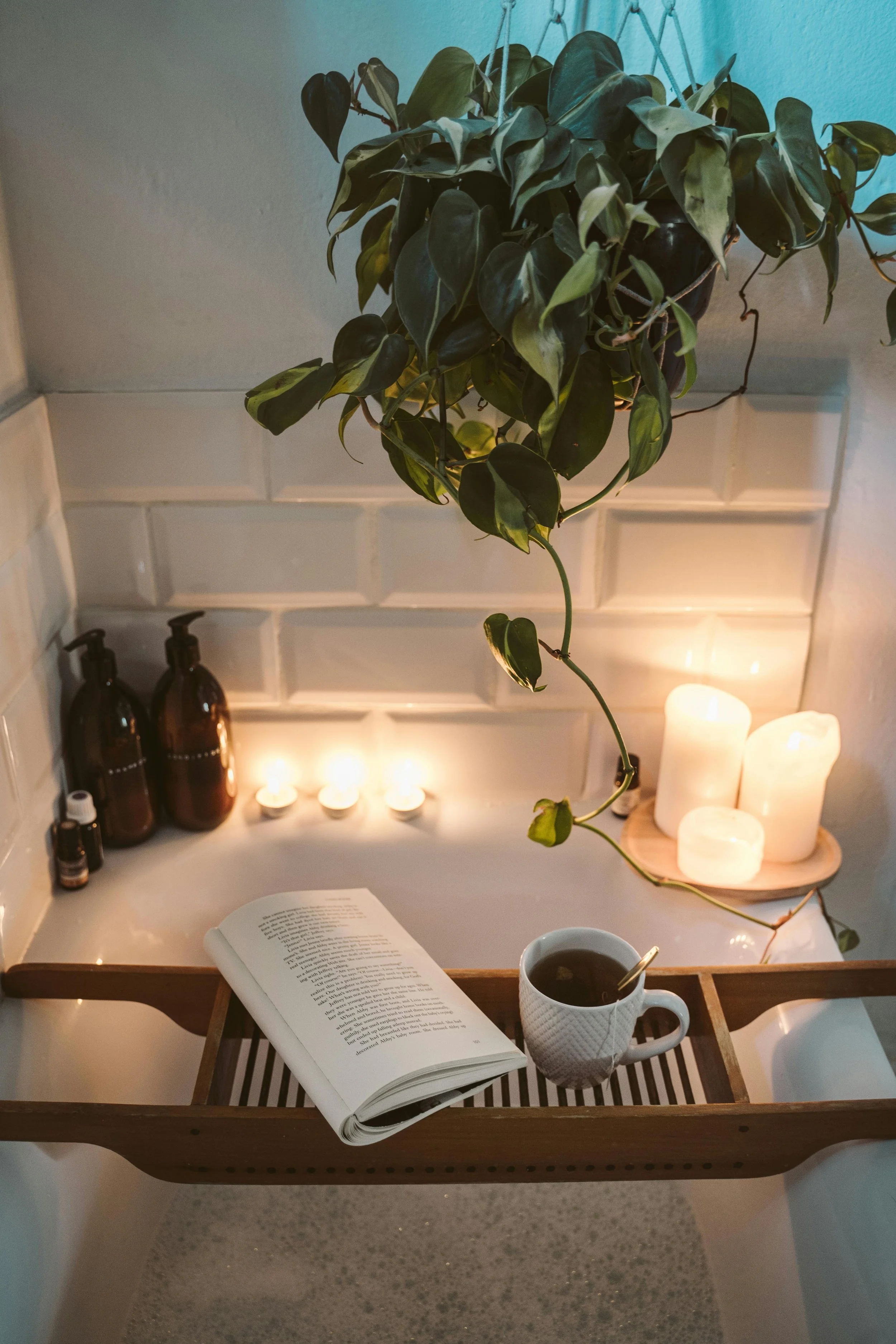 A cozy scene with a salt bath tub, a wooden bath tray holding an open book and a mug of tea, surrounded by candles and aromatherapy bottles, with a hanging green plant above.