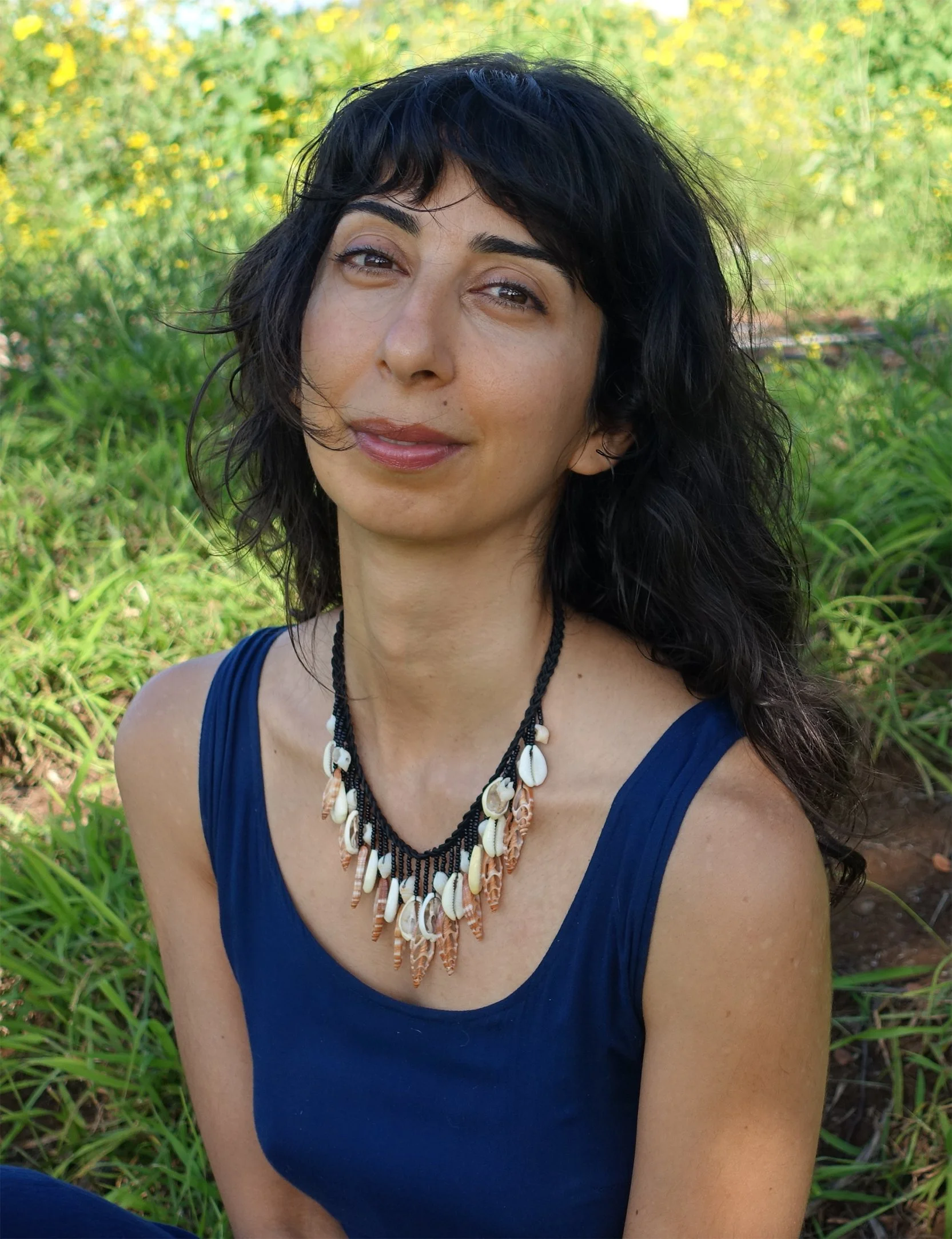 A woman with curly black hair wearing a navy blue dress and a shell necklace, smiling outdoors with green plants and yellow flowers in the background.