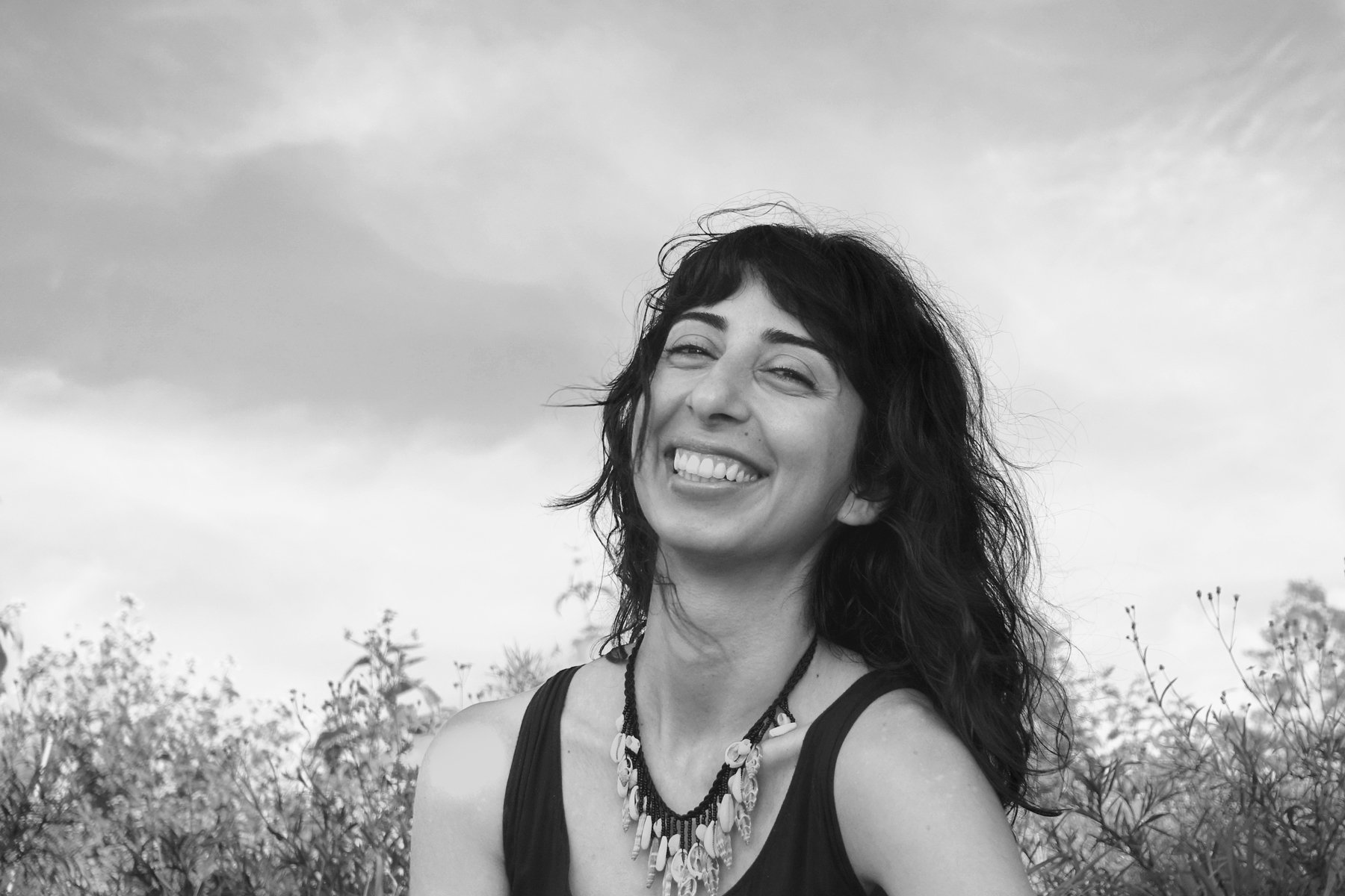 Black and white photo of a woman with long curly hair smiling outdoors, wearing a sleeveless top and a shell necklace, with trees and a cloudy sky in the background.