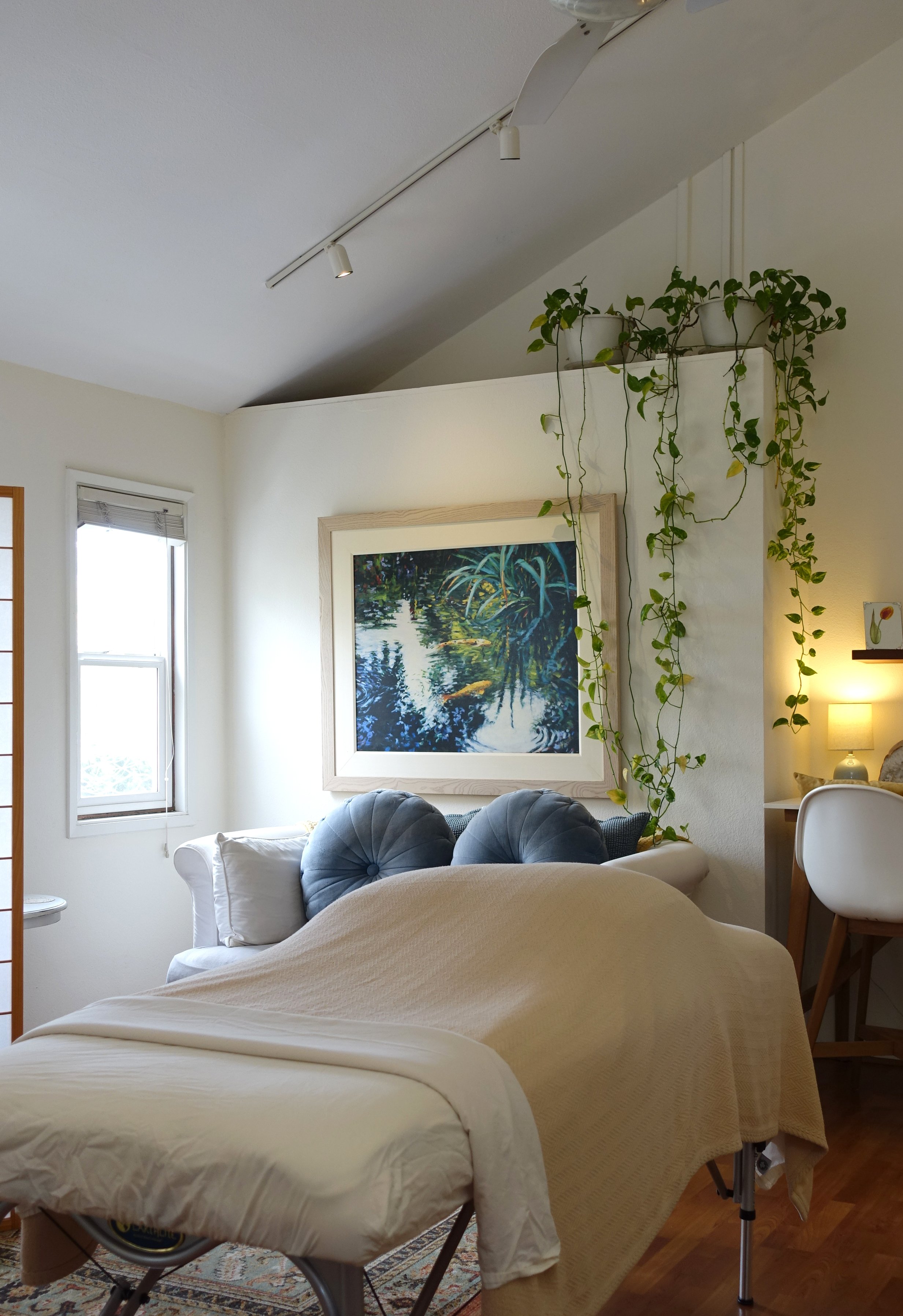 A cozy room with a white sofa, a painting of a pond with lily pads, a window, and hanging plants near the ceiling, with a massage table in the foreground.