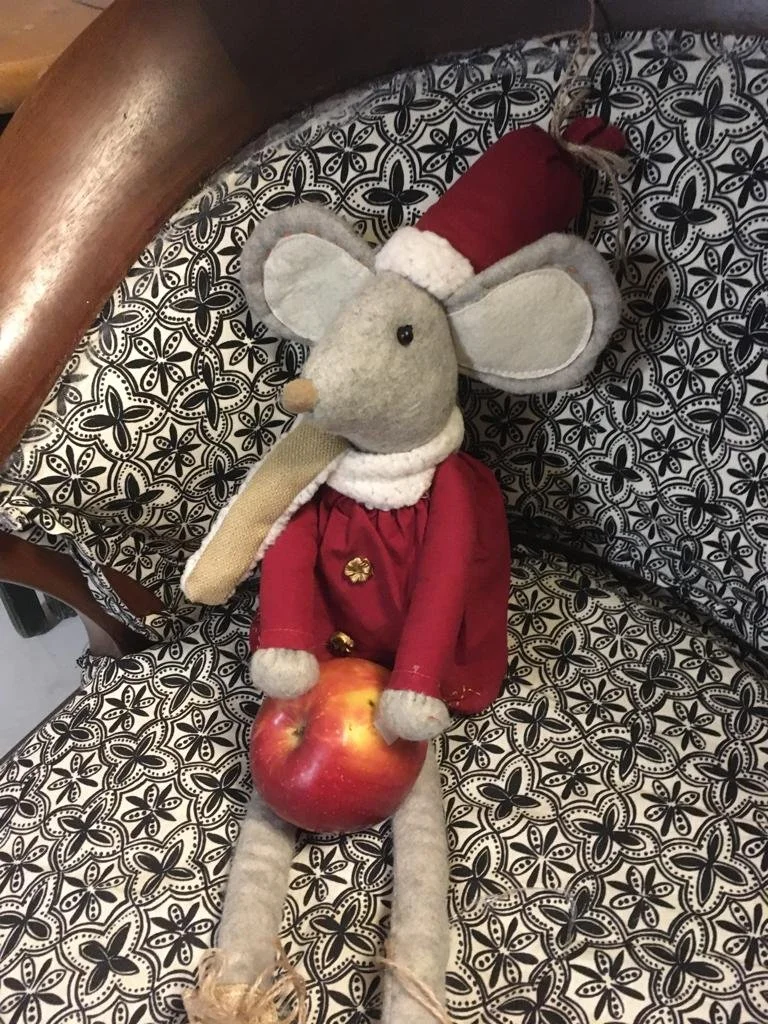 A plushSarafina mouse toy dressed in Christmas attire sitting on a patterned black and white cushion, holding an apple.