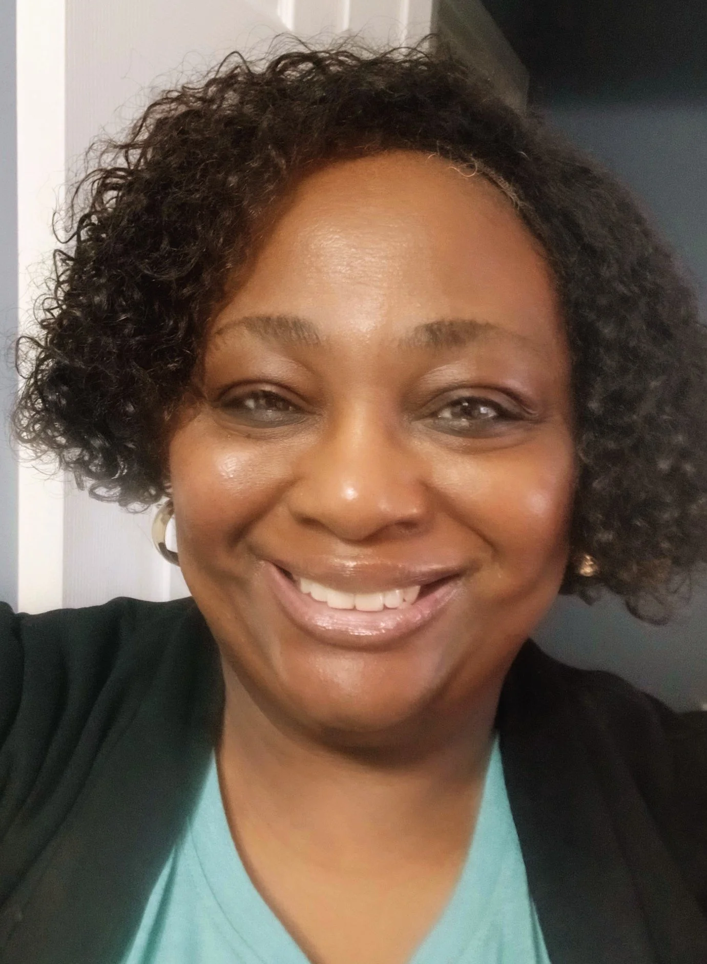 A smiling woman with curly hair, wearing a black jacket and a light turquoise top, taking a selfie in front of a gray and white background.