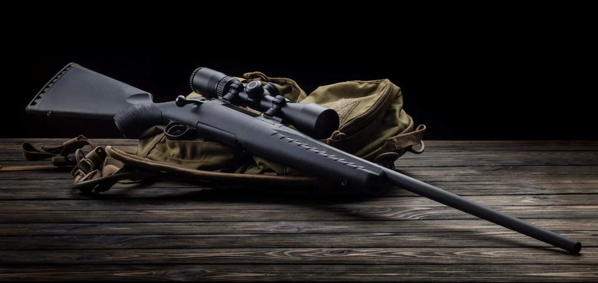A black rifle with a scope resting on an olive green backpack on a wooden surface, with a dark background.