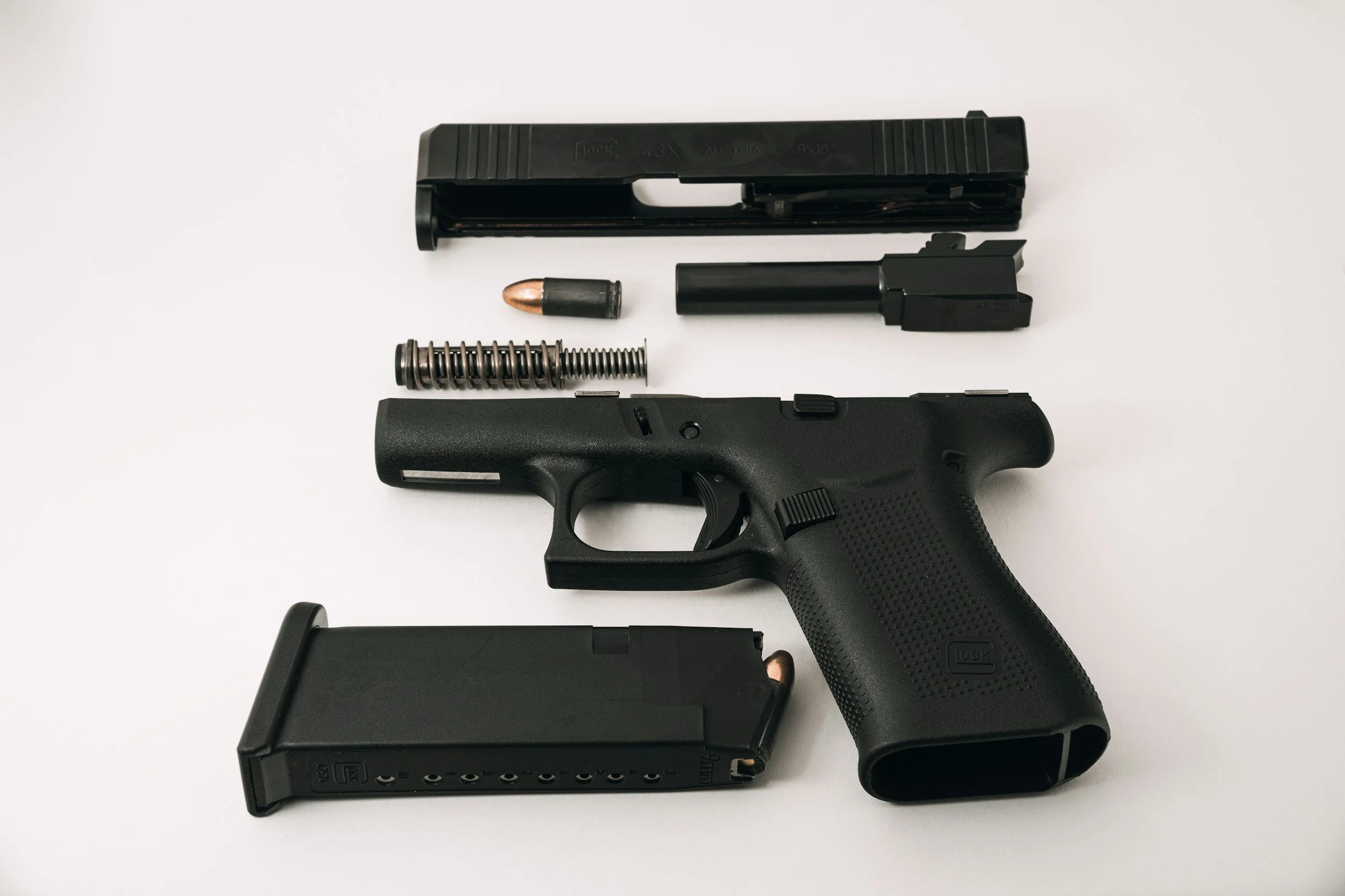 Disassembled semi-automatic handgun with magazine, slide, barrel, recoil spring, and firing pin on a white surface.