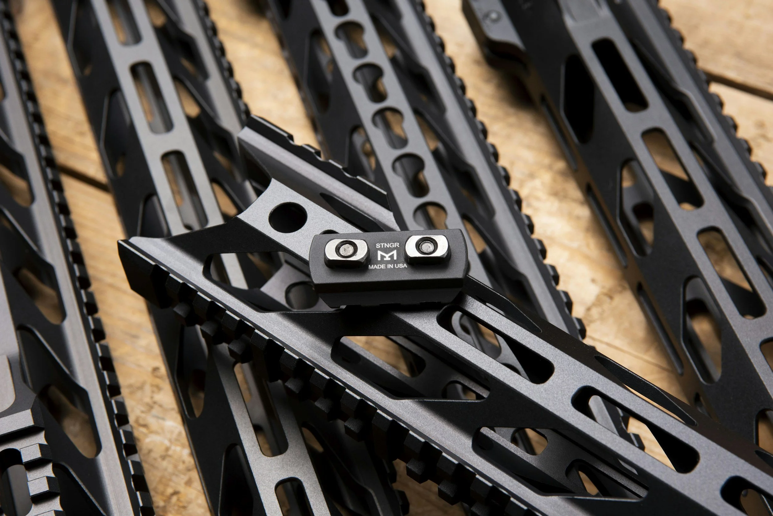 Several black rifle handguards with attachment rails are arranged on a wooden surface. One handguard features an aluminum mount labeled "STNGR M MADE IN USA."