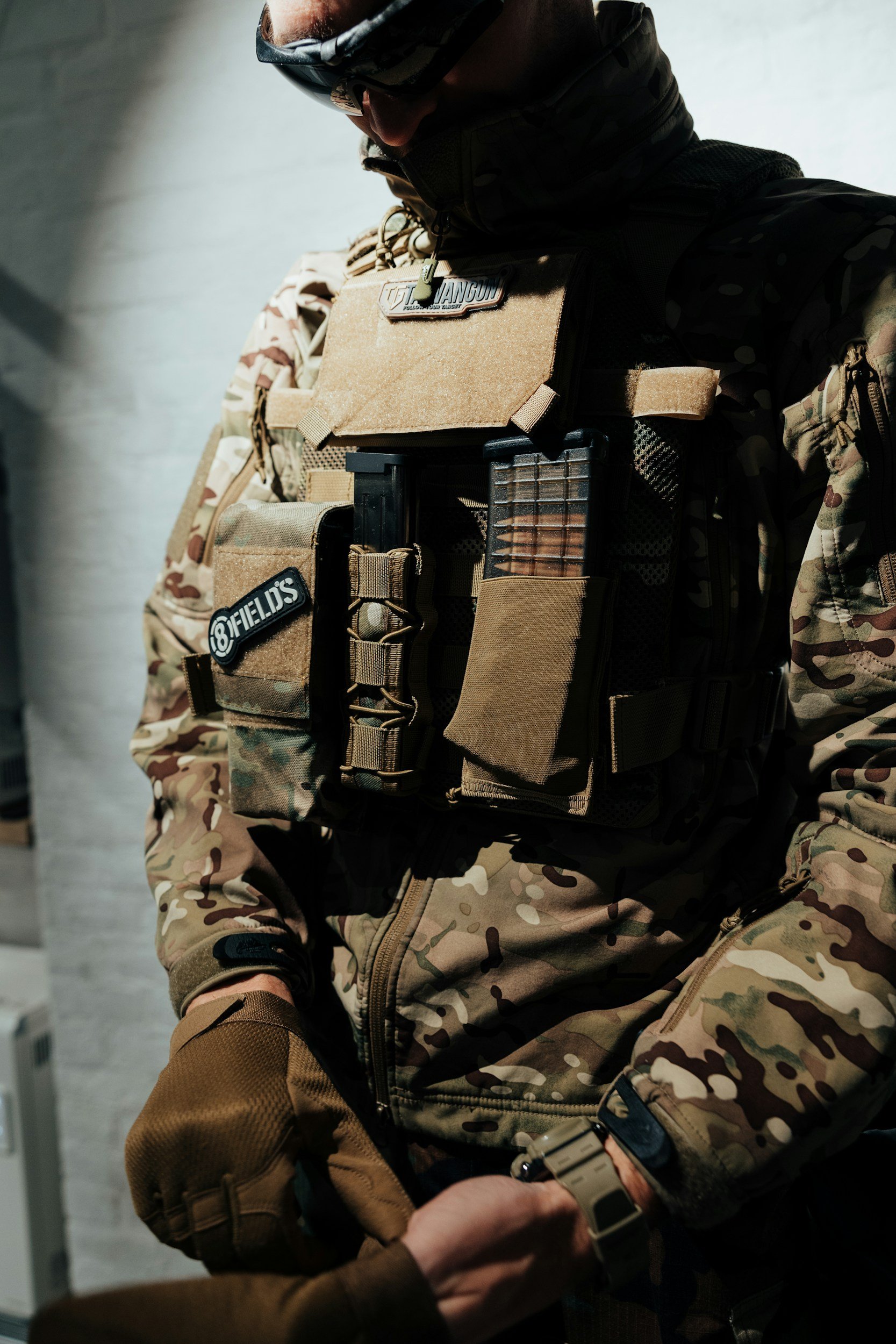 A person dressed in military tactical gear holding a handgun with a second person assisting.