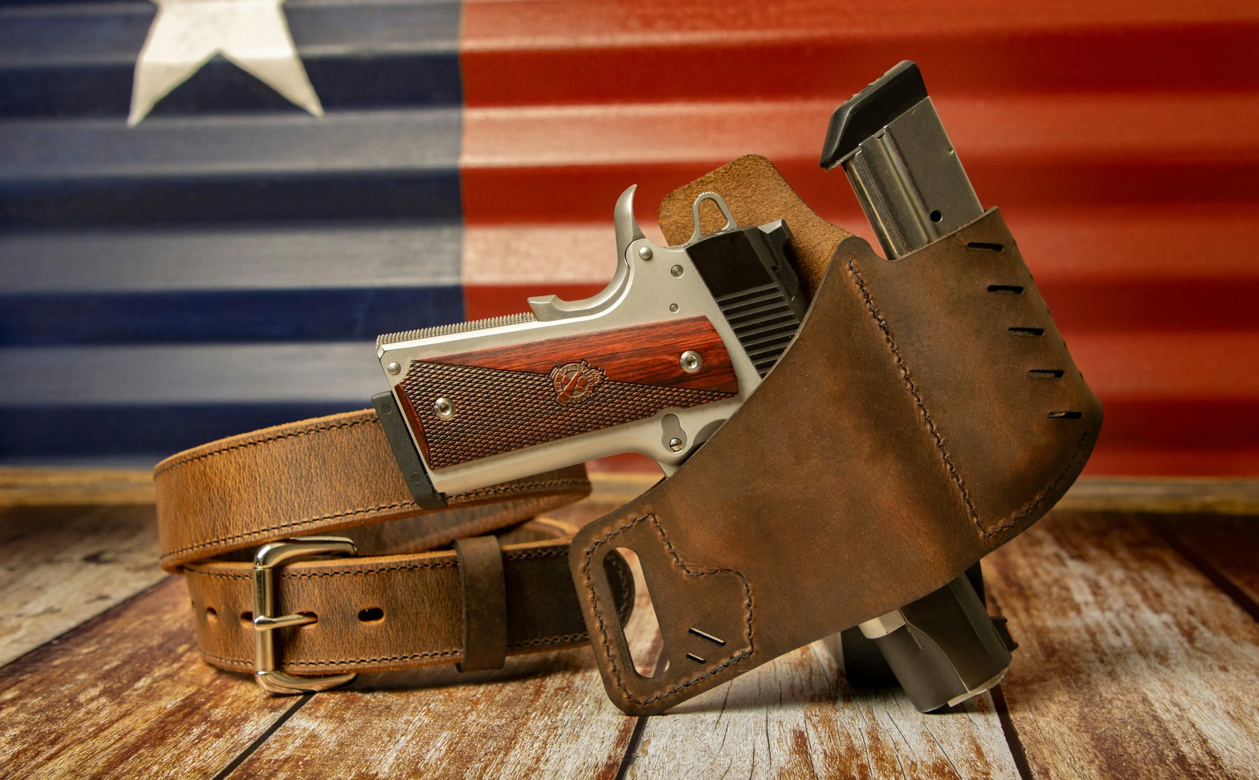 A handgun with a wooden grip and a metallic slide, resting inside a brown leather gun holster on a wooden surface. Behind them is an American flag.