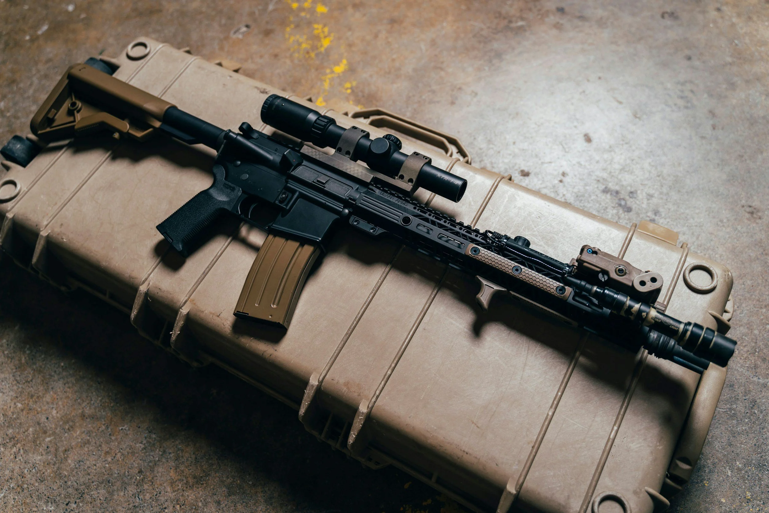 A black rifle with a scope resting on a tan hard-shell case in a concrete or industrial setting.