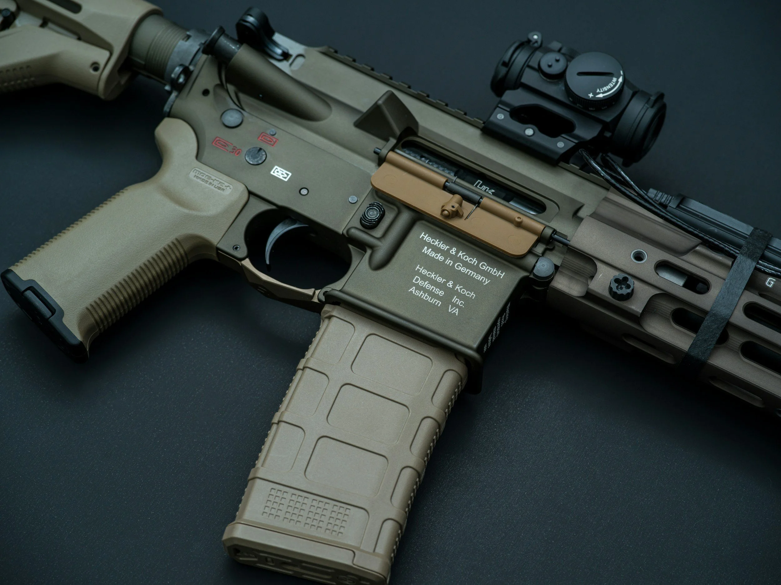 Close-up of a modern rifle with tan-colored grip and stock, with a black scope mounted on top, against a black background.