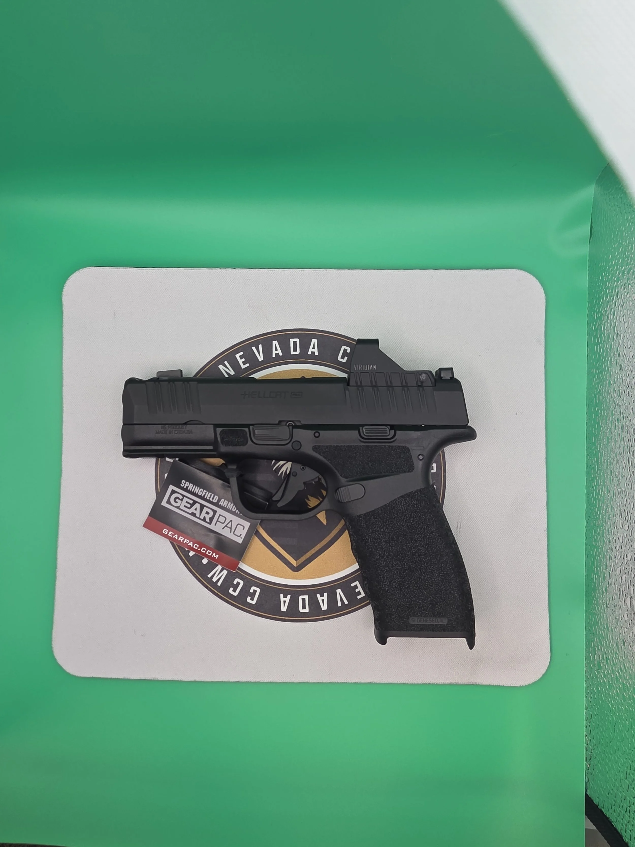 A black handgun with a textured grip resting on a white surface with a logo of Nevada on it, inside a green box.