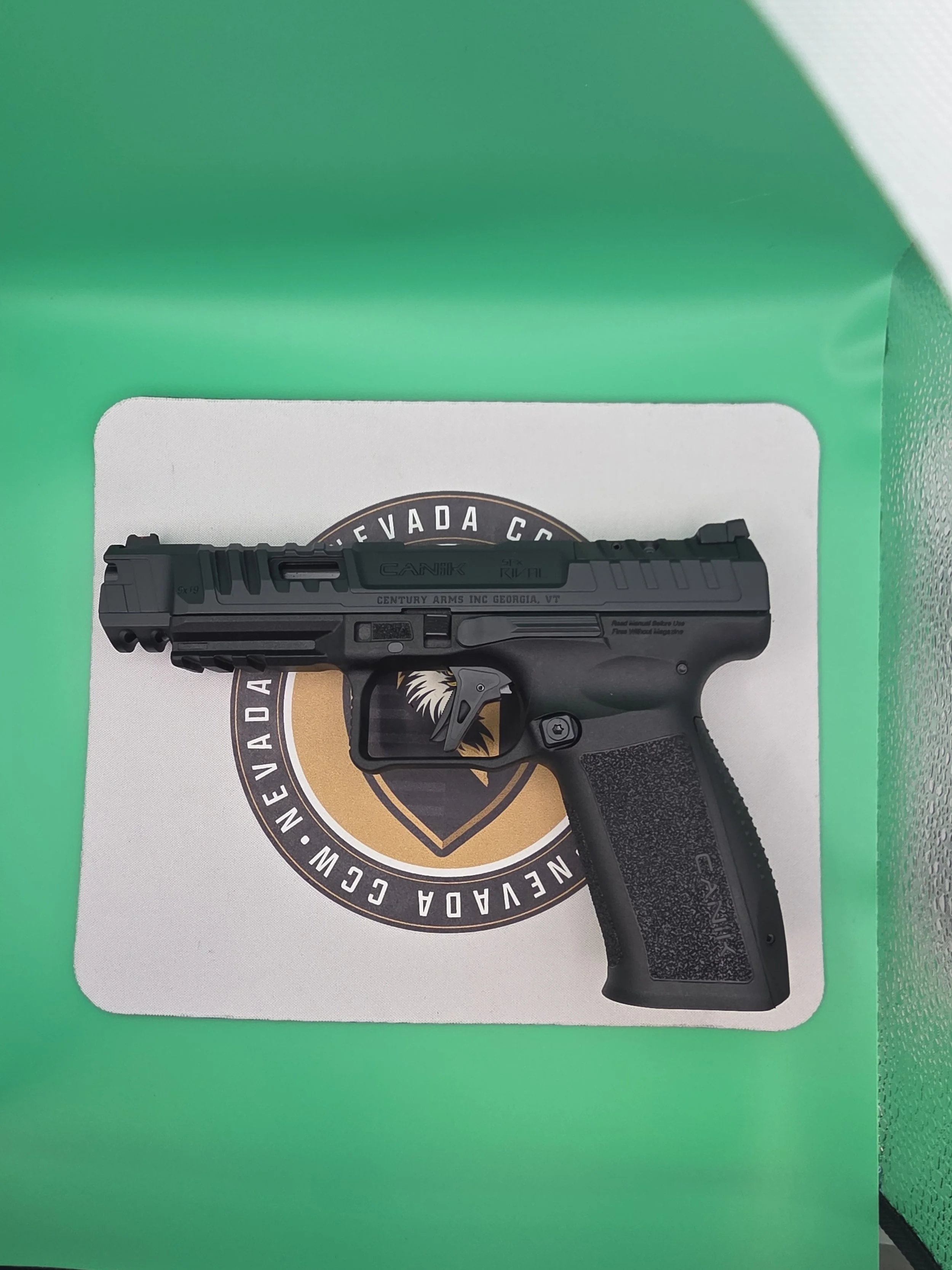 Close-up of a black handgun placed on top of Nevada Nevada CCA emblem, inside a green and white box.
