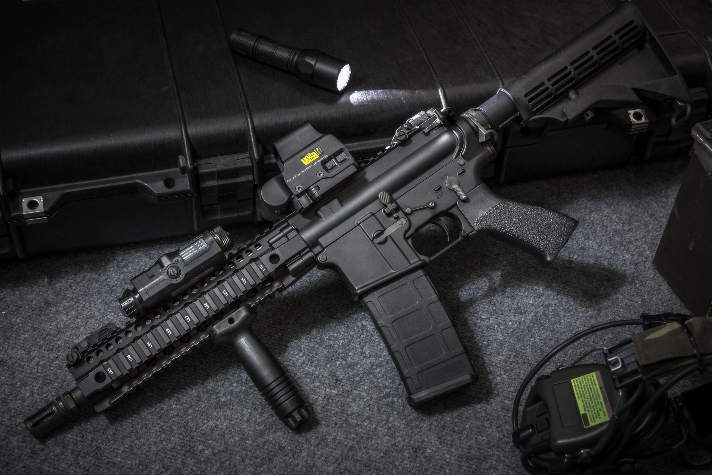 A black rifle with a bipod, mounted on a cargo box, and tactical accessories including a flashlight and sight, lying on a gray carpeted floor.