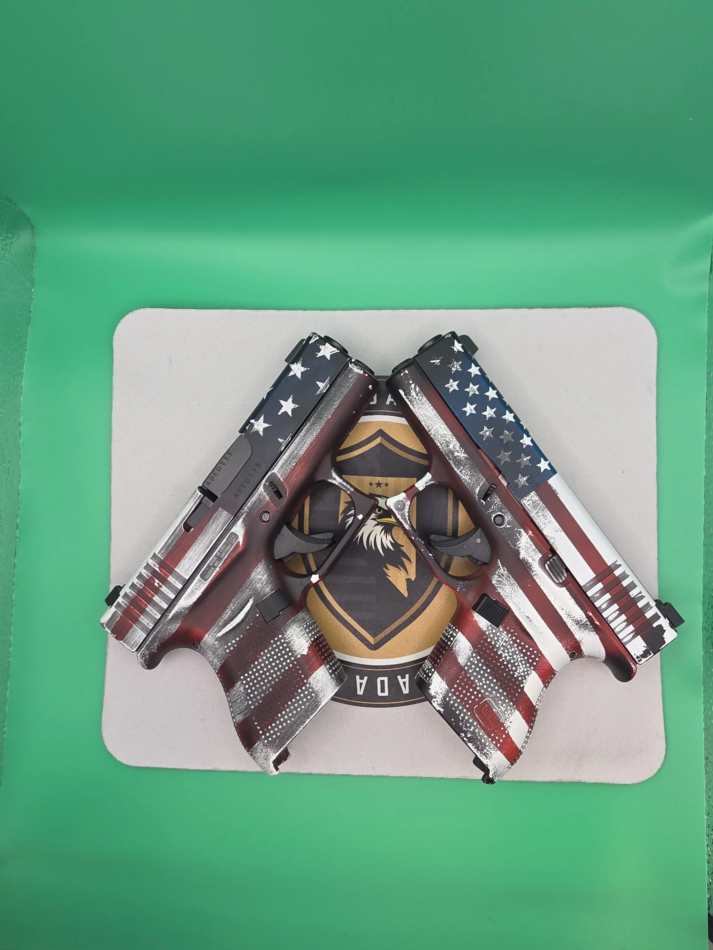 Four handguns with patriotic American flag design on their slides arranged in a circle on a emblem with a bald eagle and stars on a black background, placed on a white surface with a green background.