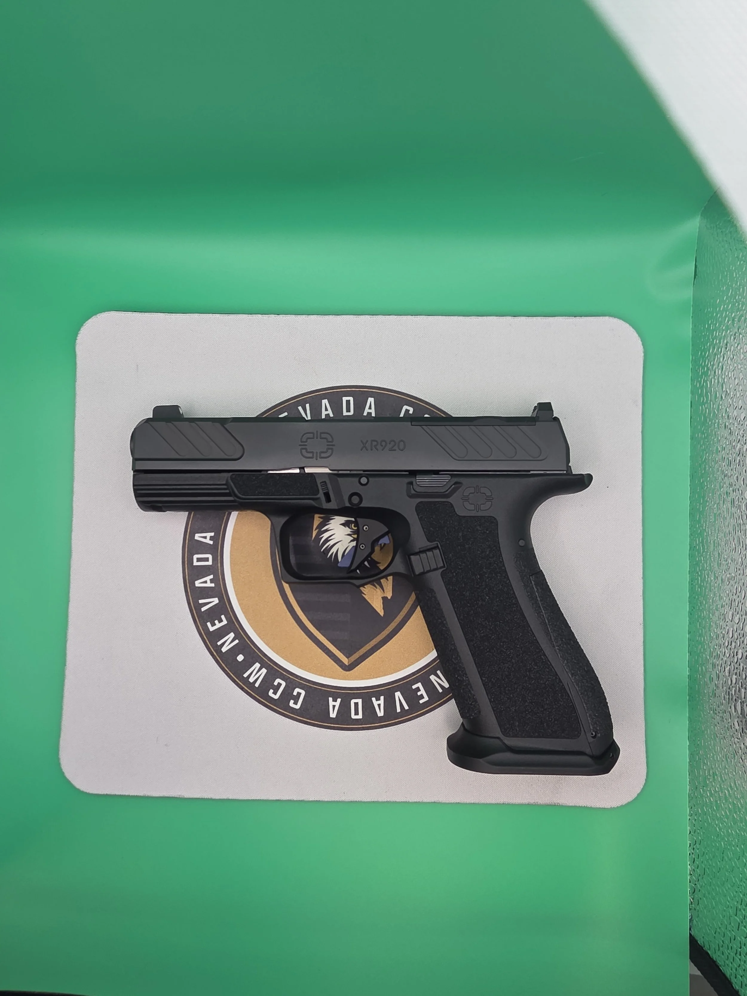 A black semi-automatic handgun with a textured grip, laid on top of a white surface with a circular logo. The logo features a lion's face in the center and a ring of text reading 'Nevada, Colorado, California, Nevada' in black and gold colors. The background is green, and part of a textured blue-green wall is visible on the right edge.