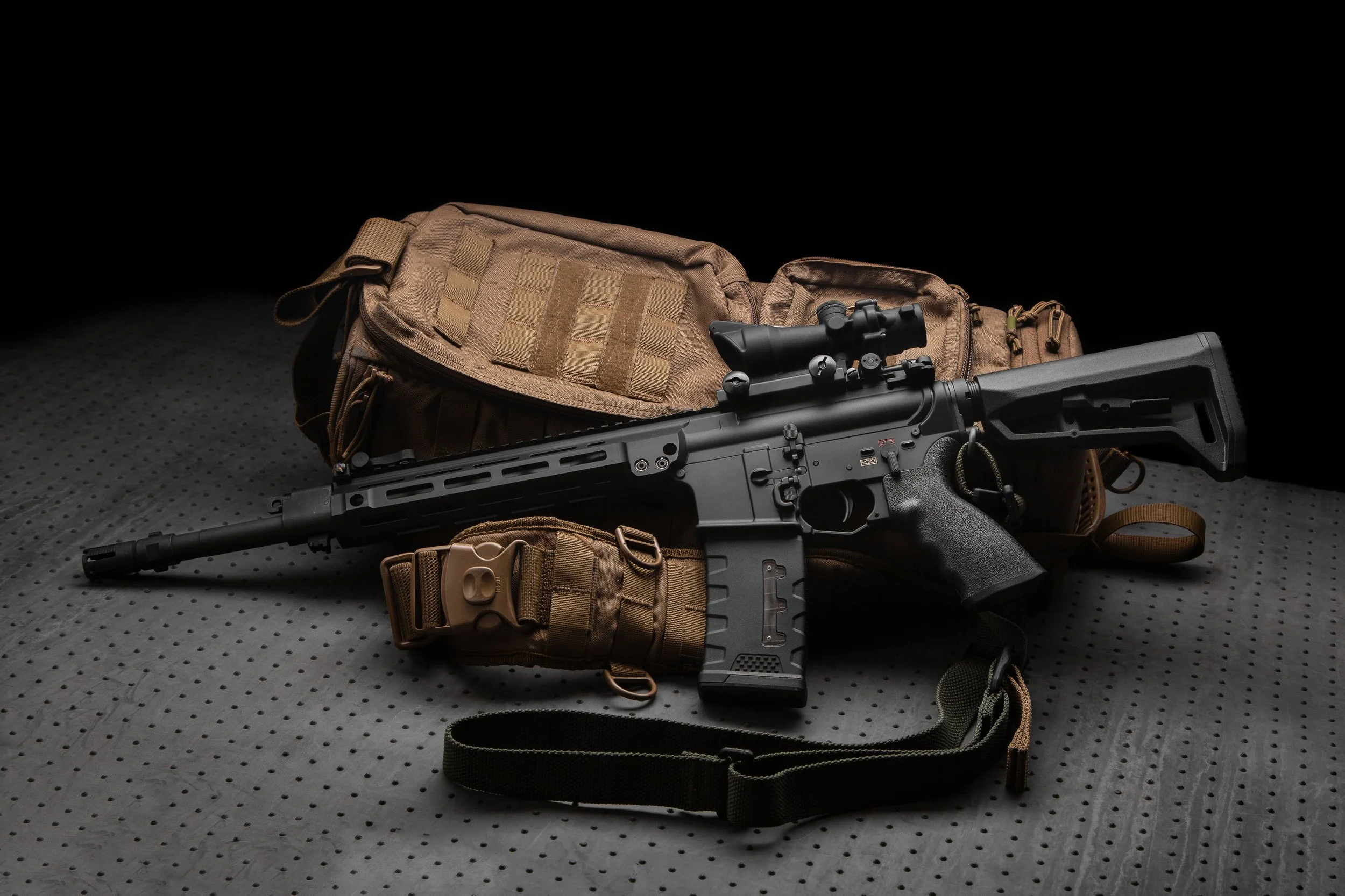 Military gear including a black rifle with scope, tan tactical backpack, pouches, and straps on a dark textured surface.