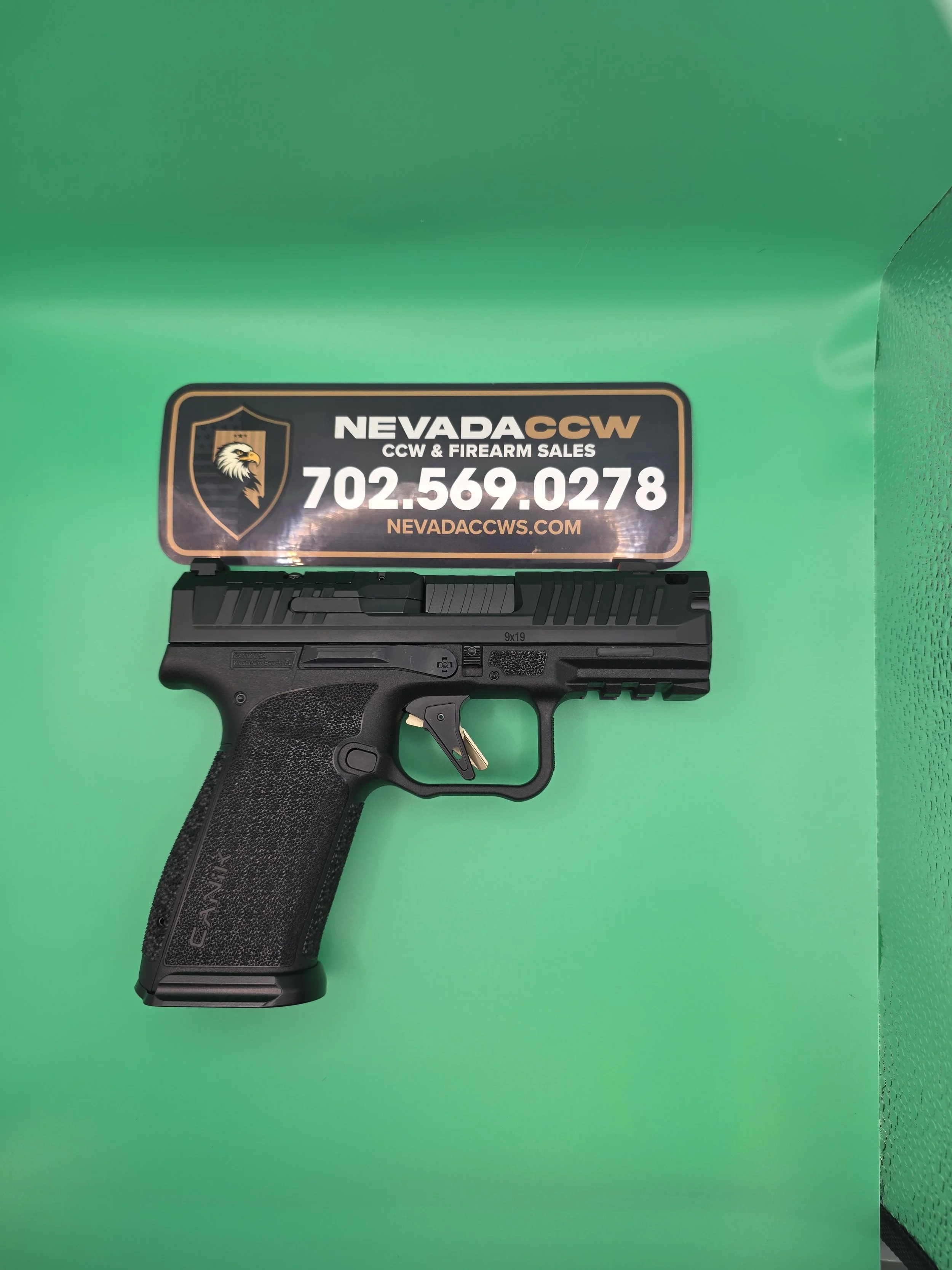 A black handgun with textured grip, partially visible, placed on a green background. A sticker with a shield, an eagle logo, and contact information for Nevada CCW and firearm sales is above it.
