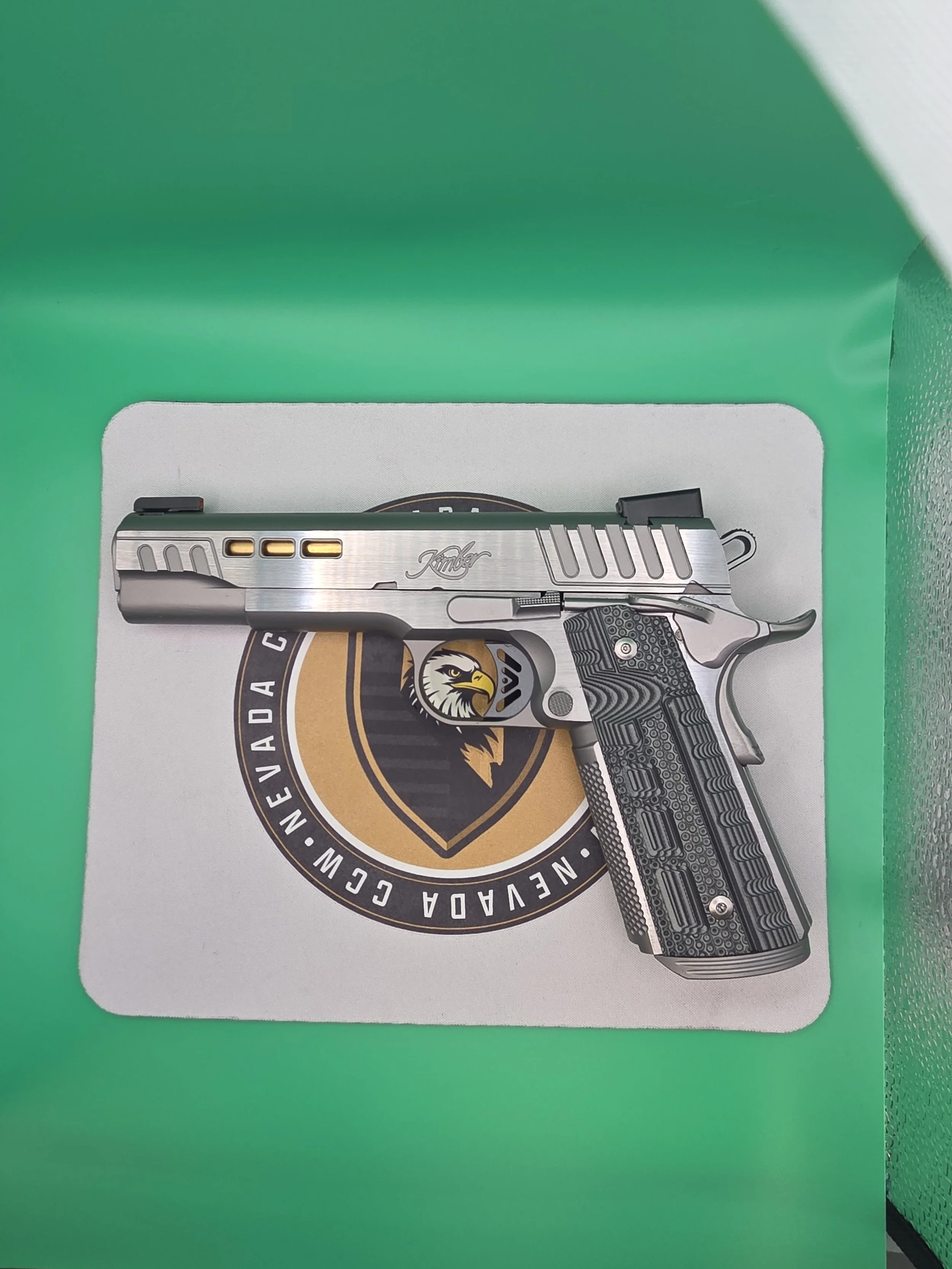 A cardboard box with a green top and a white bottom featuring a graphic of a silver and black handgun with a textured grip, set against a green background.