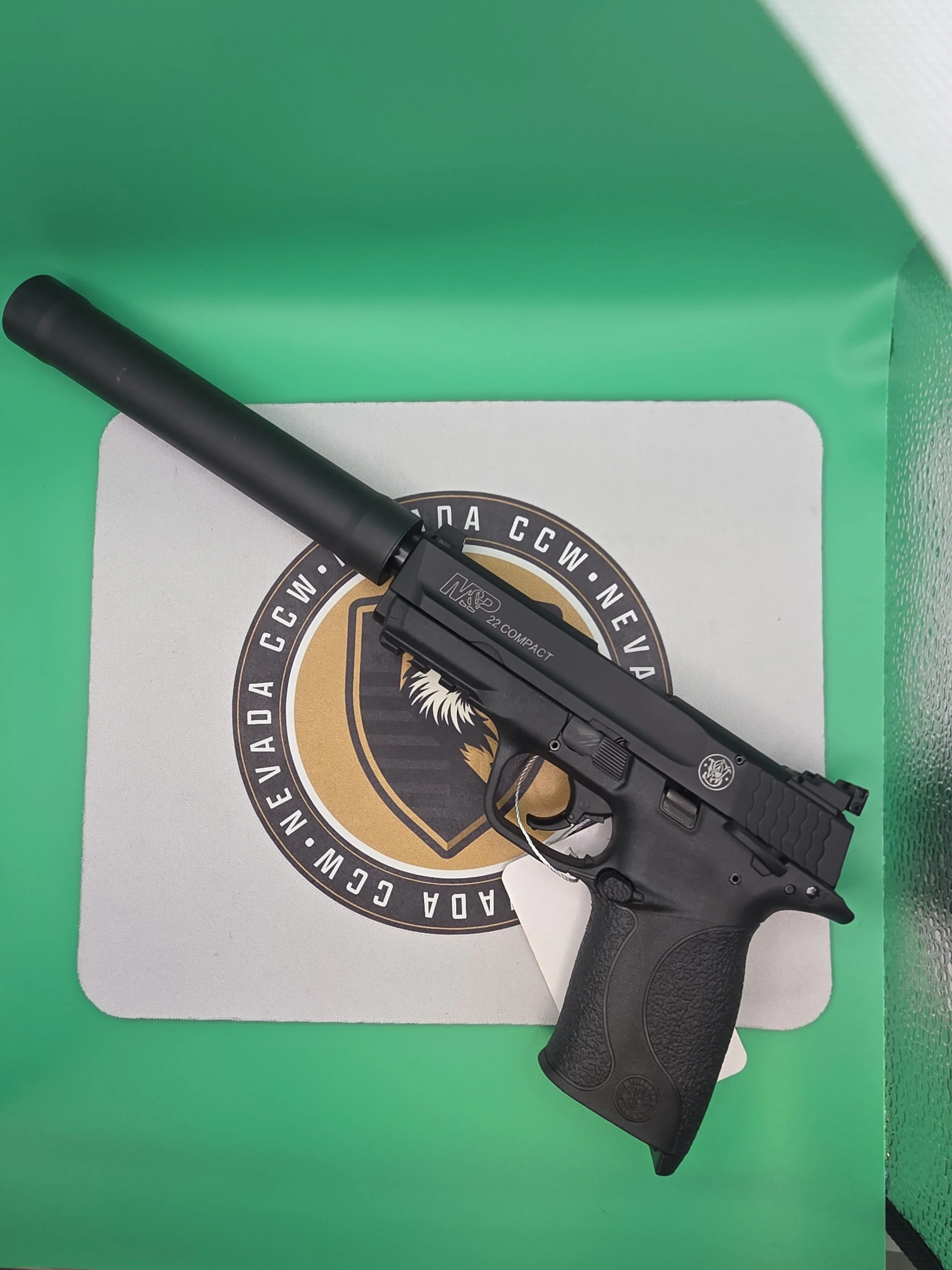 A black handgun with a silencer attached, resting on a box with a logo and text, on a white and green surface.