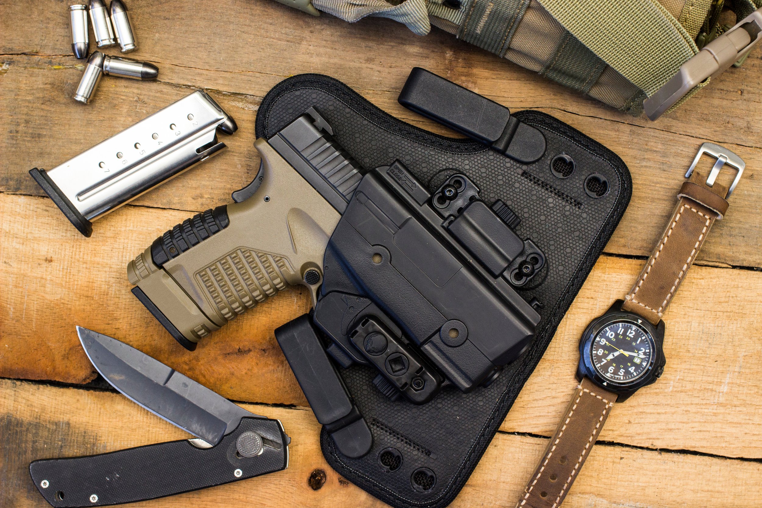 A table with a handgun, magazine, knife, flashlight, watch, and tactical gear, all arranged on a wooden surface.