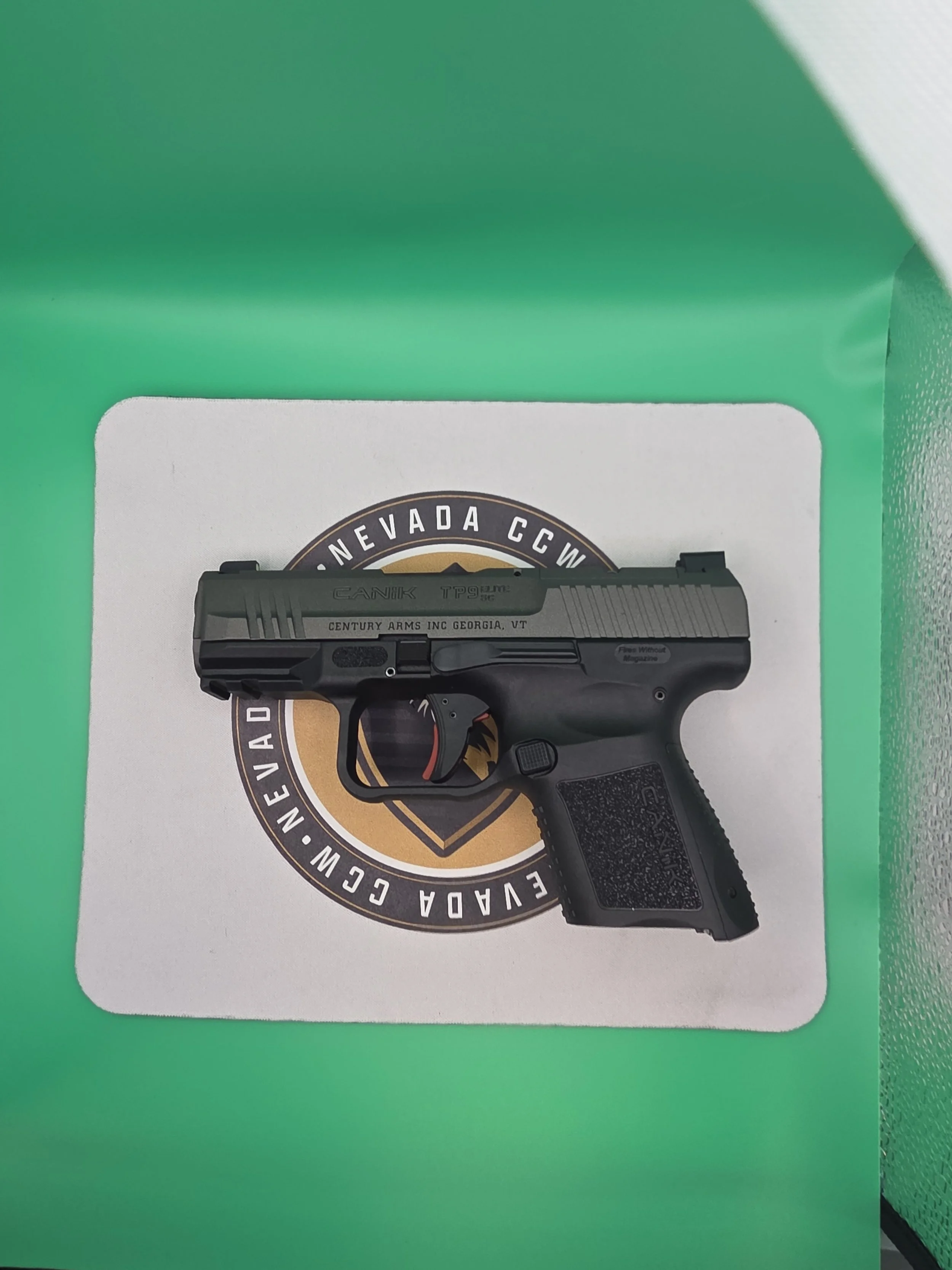 Photograph of a black semi-automatic handgun with markings, placed over a paper with a logo and text, on a green background.