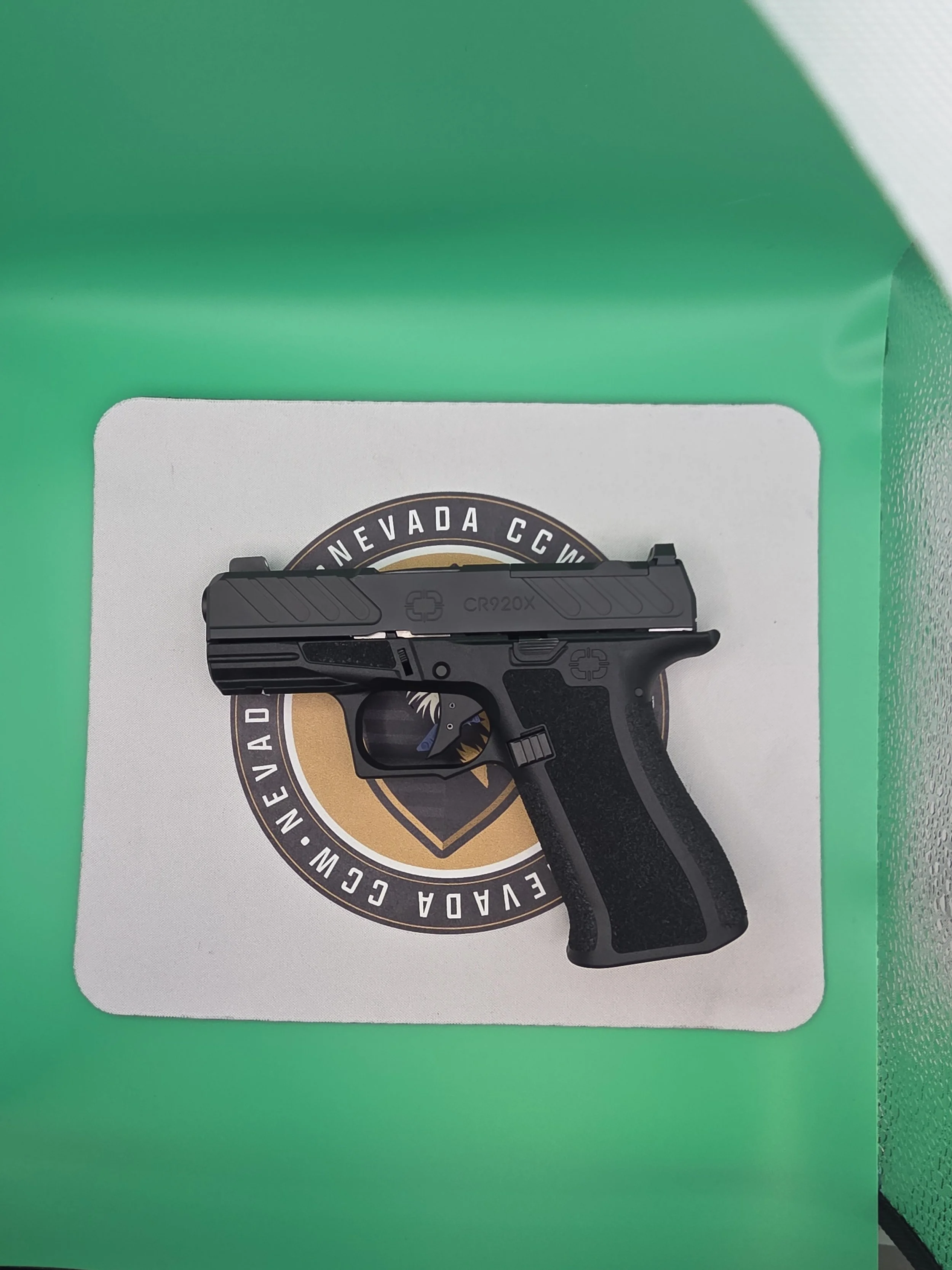 A black handgun with a textured grip and slide resting on a white square surface with a circular emblem, against a bright green background.