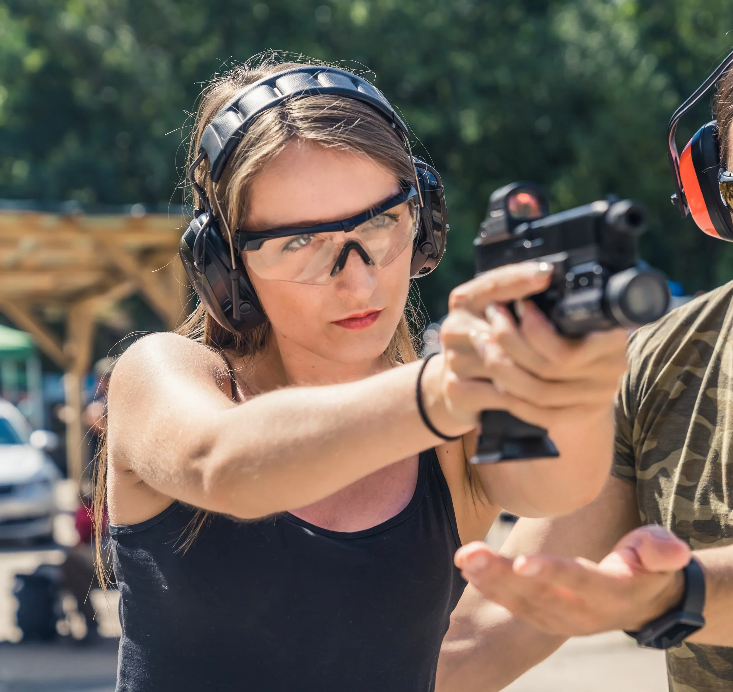 A young woman with light brown hair wearing safety goggles and a helmet, aiming a handgun outdoors with greenery and blurred background.